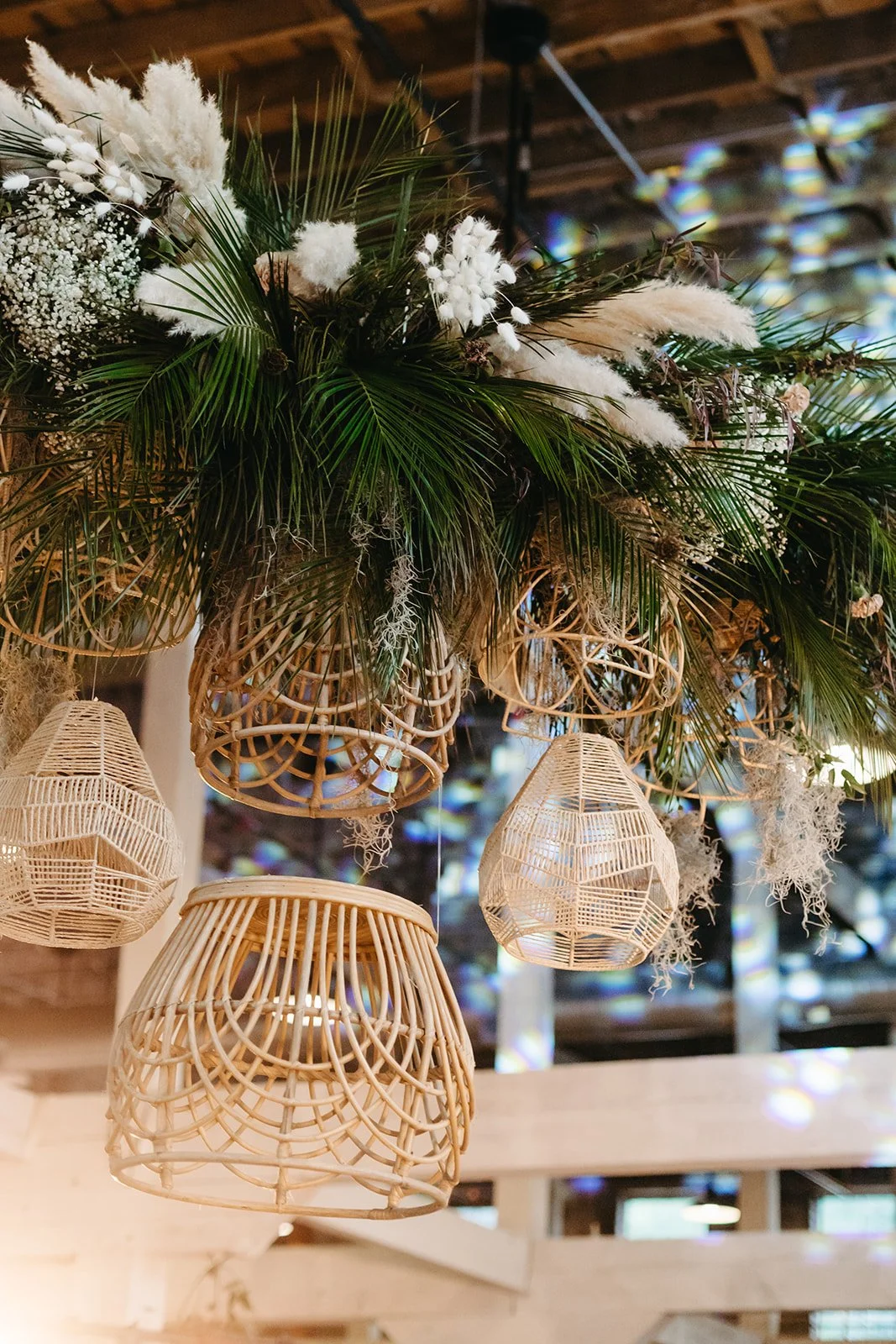 Decorative hanging wicker light fixtures and a floral arrangement with palm leaves and white flowers, hanging from a ceiling inside a rustic building.