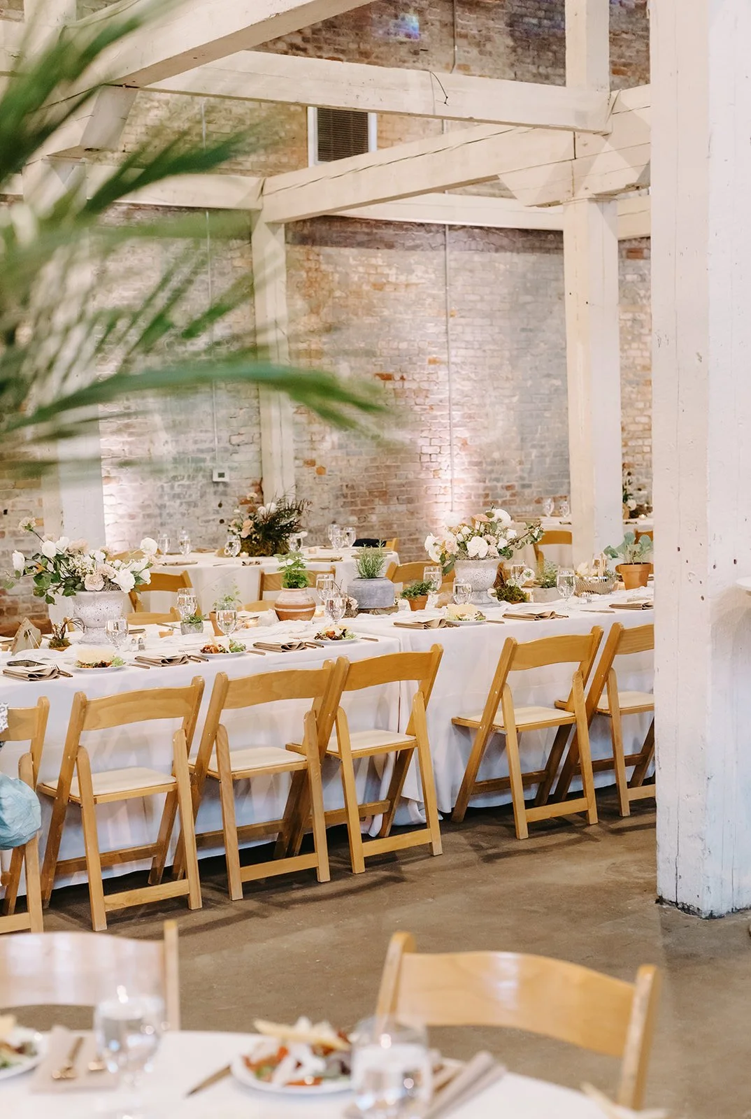 A banquet table decorated with white tablecloths, floral centerpieces, and glassware, set in an industrial-style venue with exposed brick walls and wooden beams.