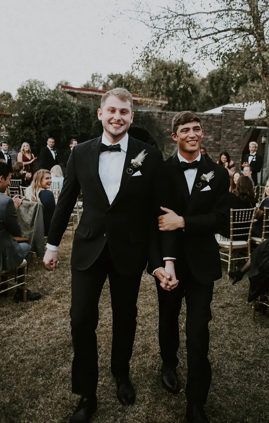 Two men in black tuxedos walking arm in arm at an outdoor wedding ceremony, surrounded by seated guests and standing wedding party, with trees and a stone building in the background.