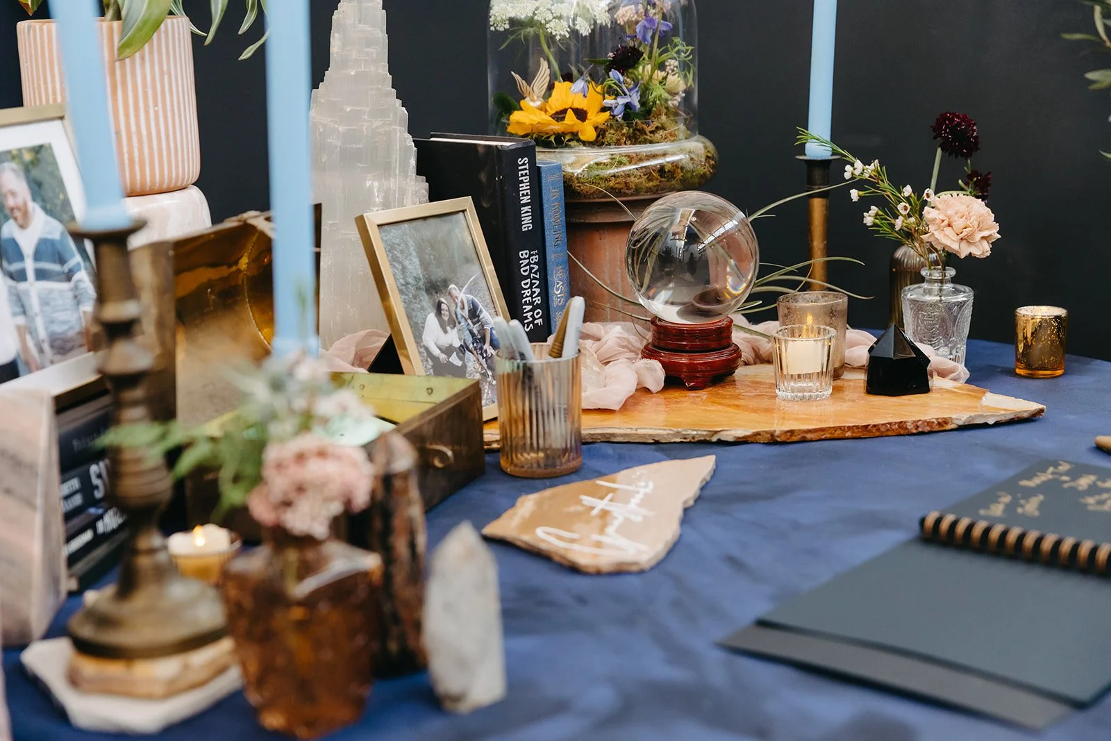 A table decorated with flowers, candles, framed photographs, books, and decorative items for a special occasion, with a dark blue tablecloth and a wooden centerpiece.