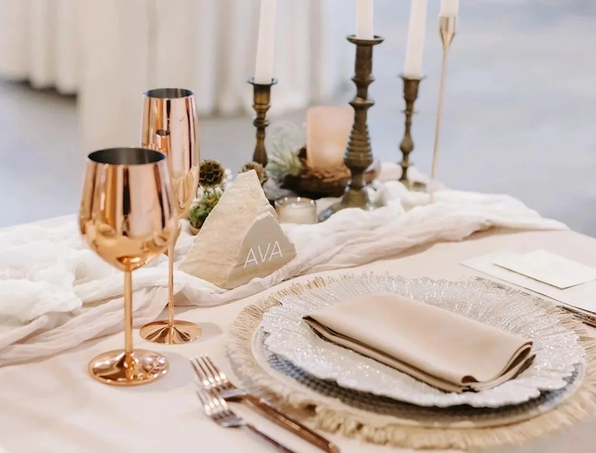 Elegant table setting with rose gold wine glasses, beige napkins, and decorative candles on a cream-colored tablecloth.