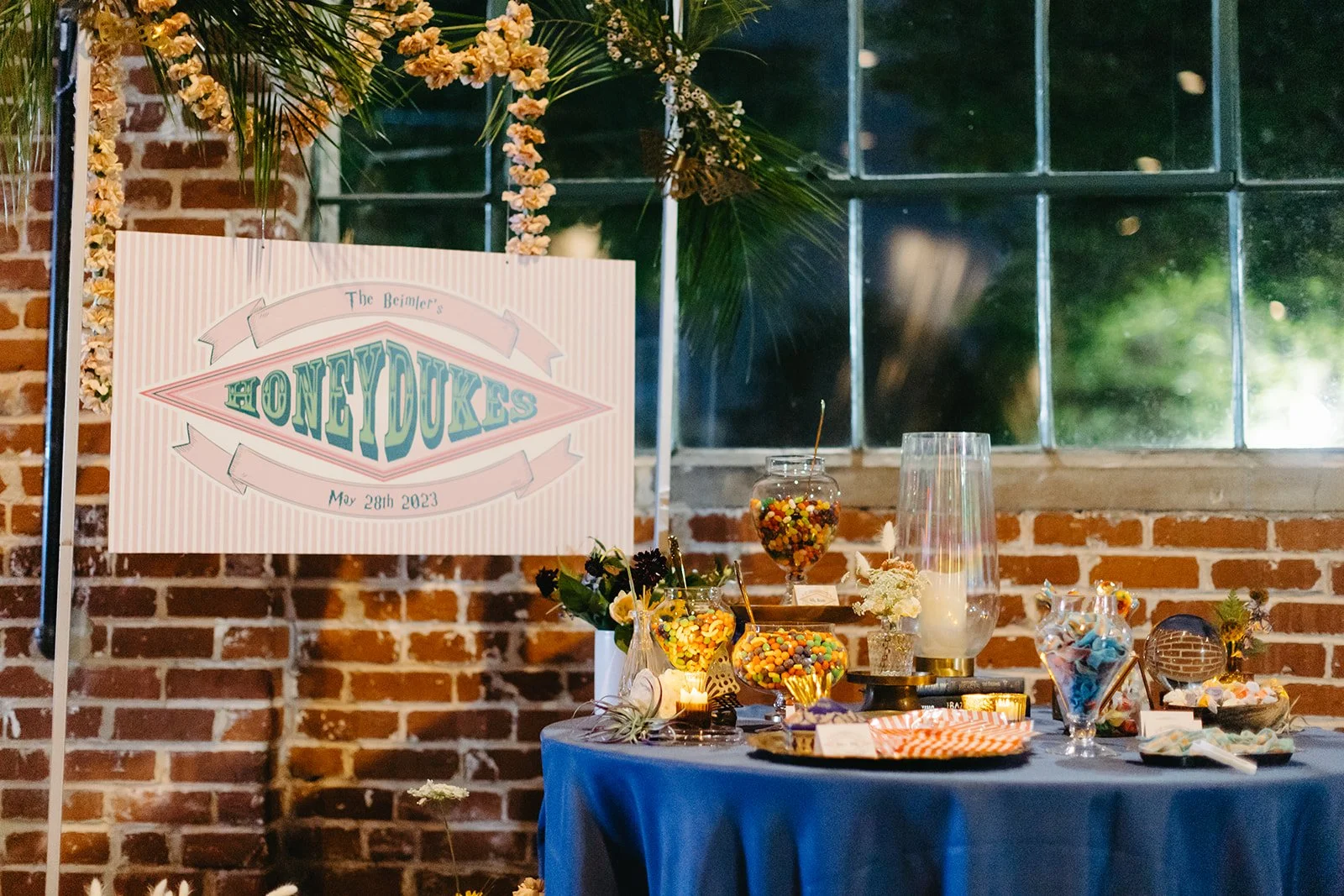 Dessert table with candy jars, flowers, and a large sign that reads "The Beimler's Honeydukes, May 28th, 2023". The table has a blue tablecloth and is set against a brick wall with a window in the background decorated with greenery and flowers.
