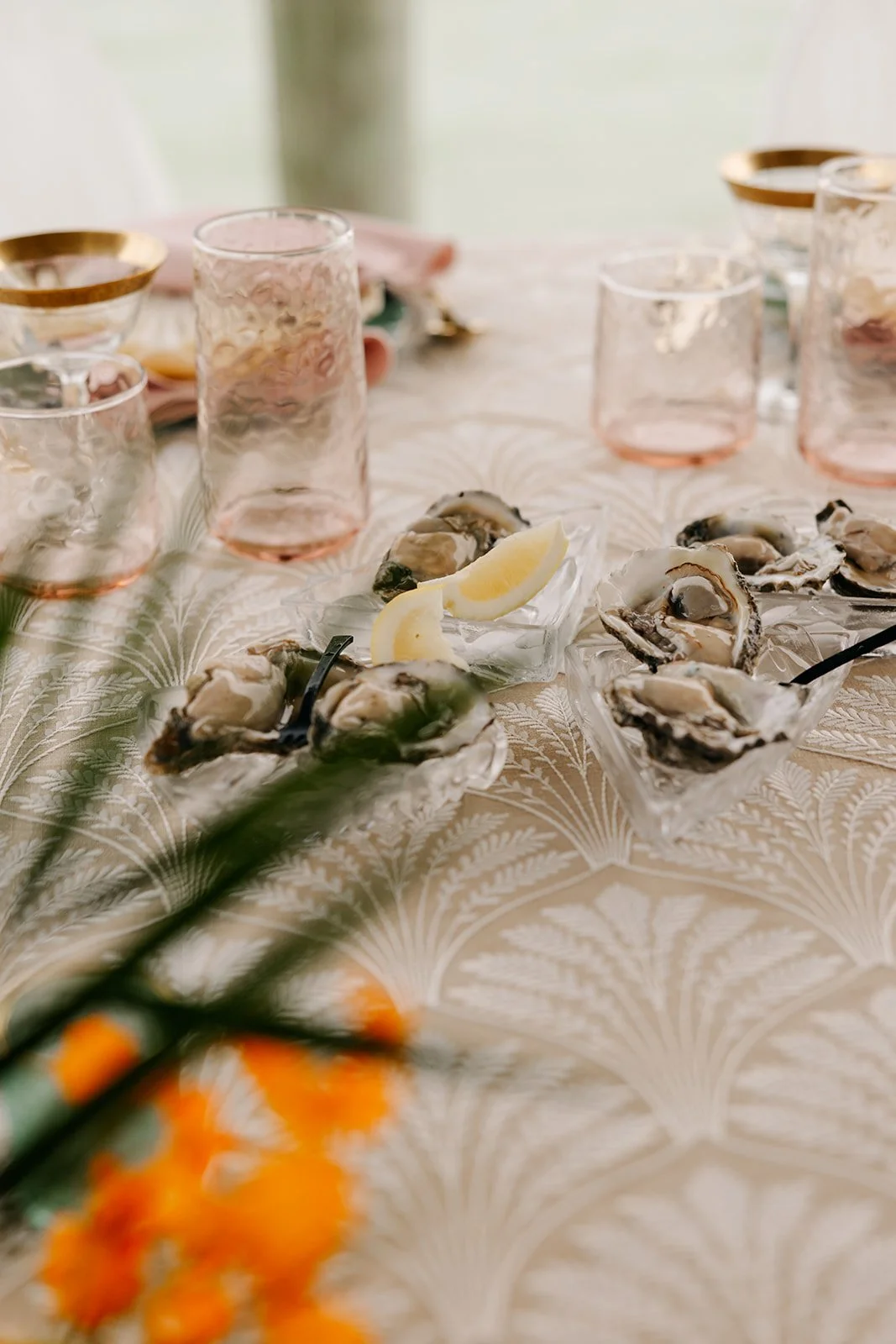 A table set with oysters on the half shell, lemon wedges, pink drink glasses with gold rims, and a floral tablecloth, with blurred orange flowers in the foreground.