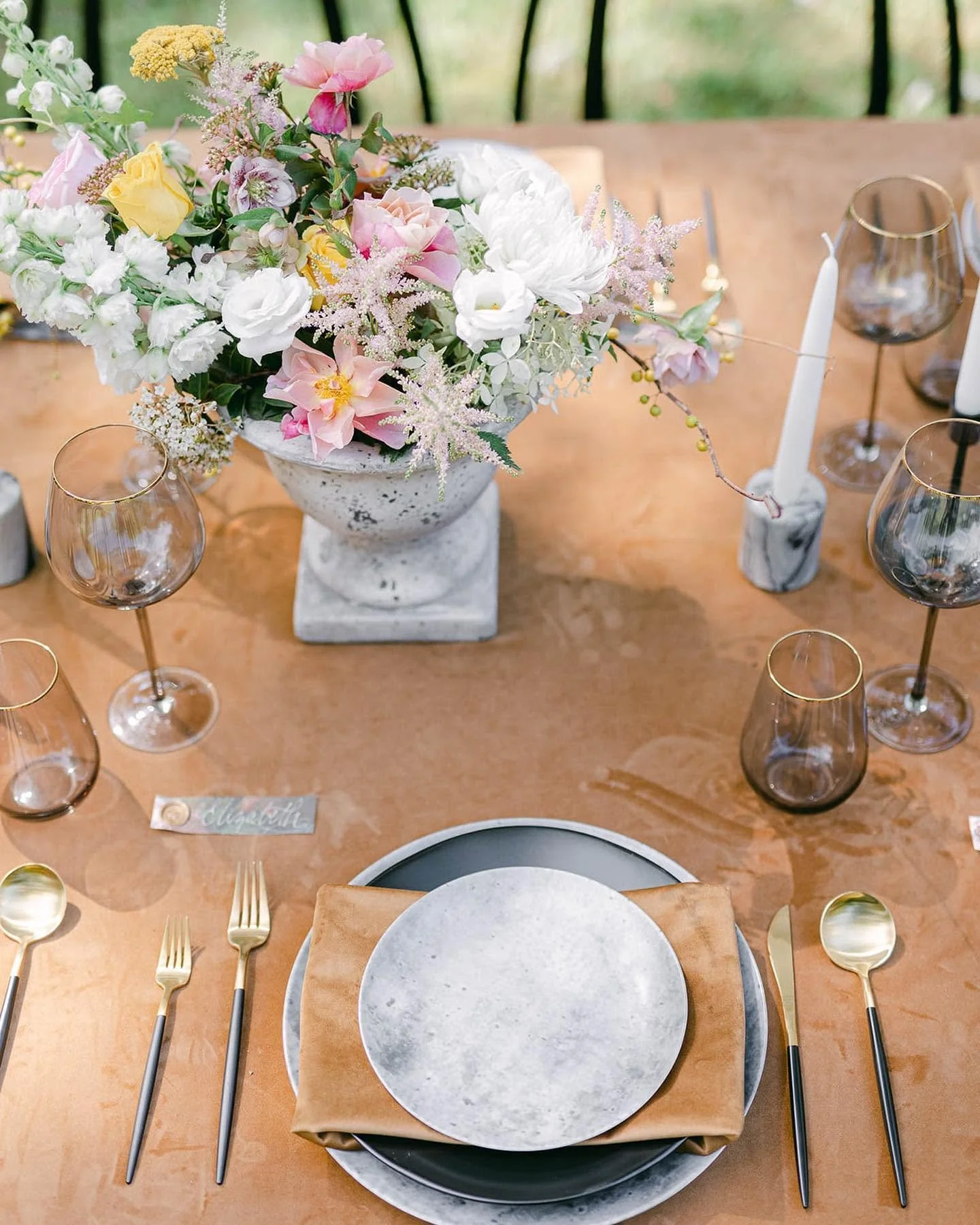 Elegant table setting with a floral centerpiece, gold flatware, dark and clear glasses, a place card, and a marble plate on a wooden table.