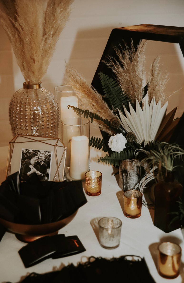 Decorative table with candles, a framed black-and-white photo of two people, a large beaded vase with dried pampas grass, a mirror, and various plants in vases.