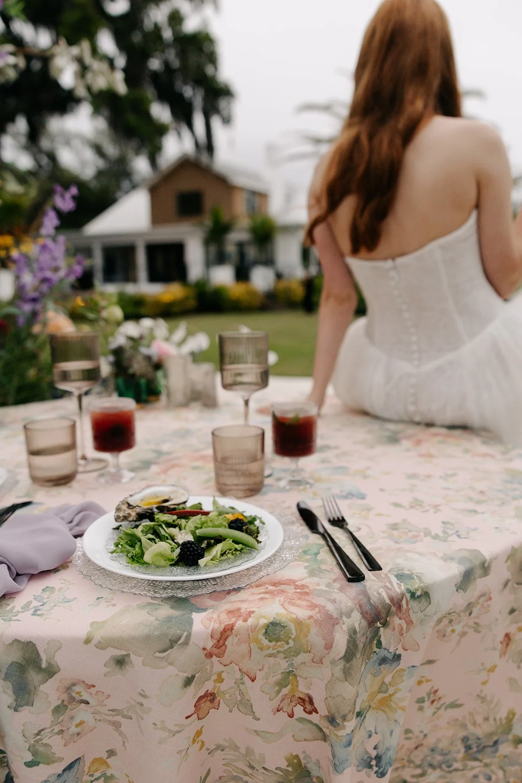 A table set for a meal with a scenic backyard view, featuring a salad plate with greens and blackberries, glasses with drinks, and utensils, with a woman in a white dress sitting on the table.