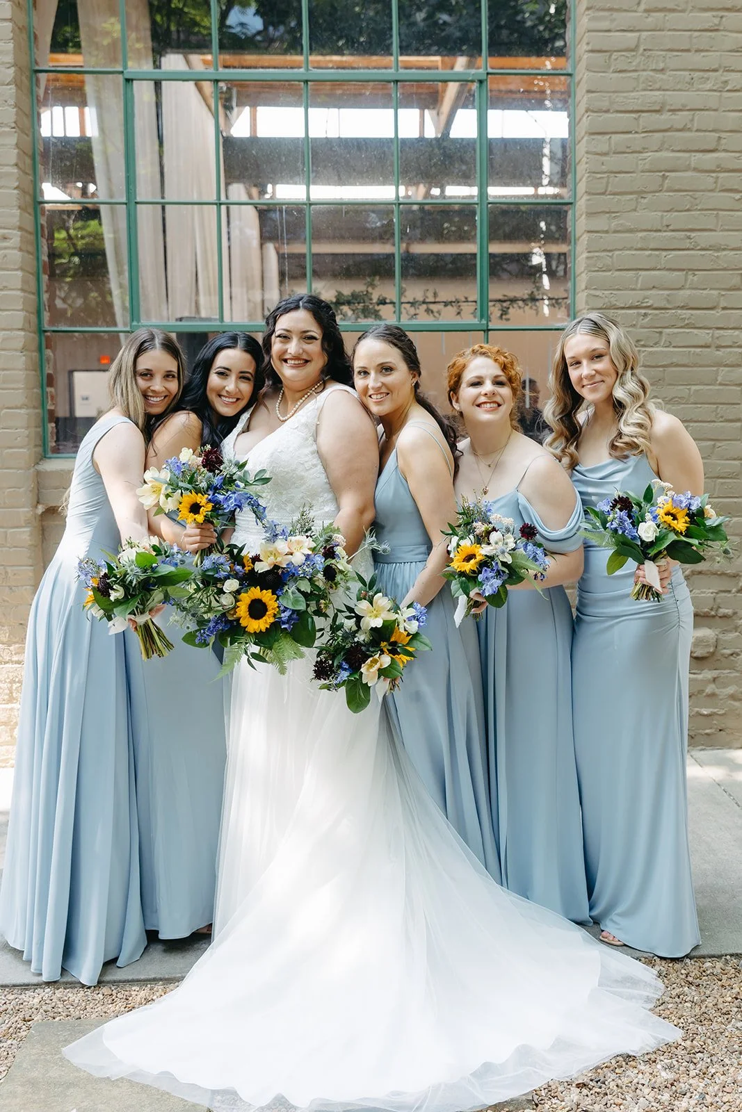 A bride and five bridesmaids standing together, smiling, dressed in light blue gowns and holding colorful bouquets of flowers, in front of a brick wall with a large window.