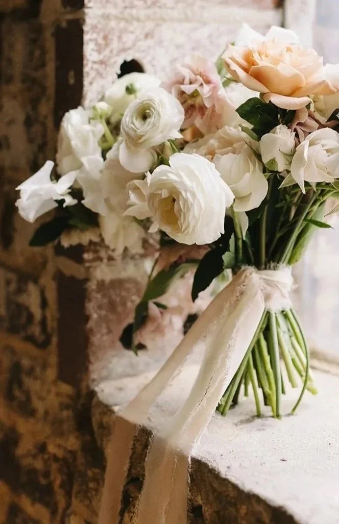 A bouquet of white and soft pink flowers tied with a white ribbon, placed on a rustic brick ledge.