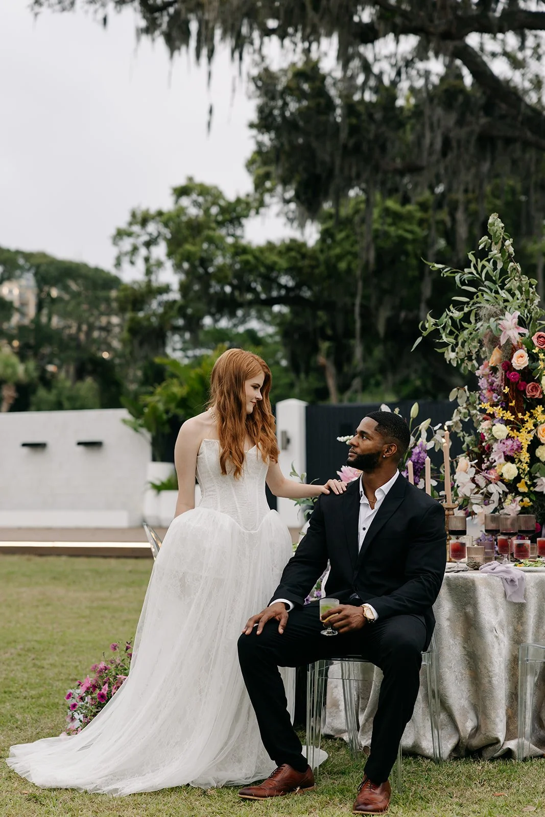 A bride with long red hair in a white wedding dress stands next to a seated groom in a black suit at an outdoor wedding reception, with a decorated table and floral arrangements.