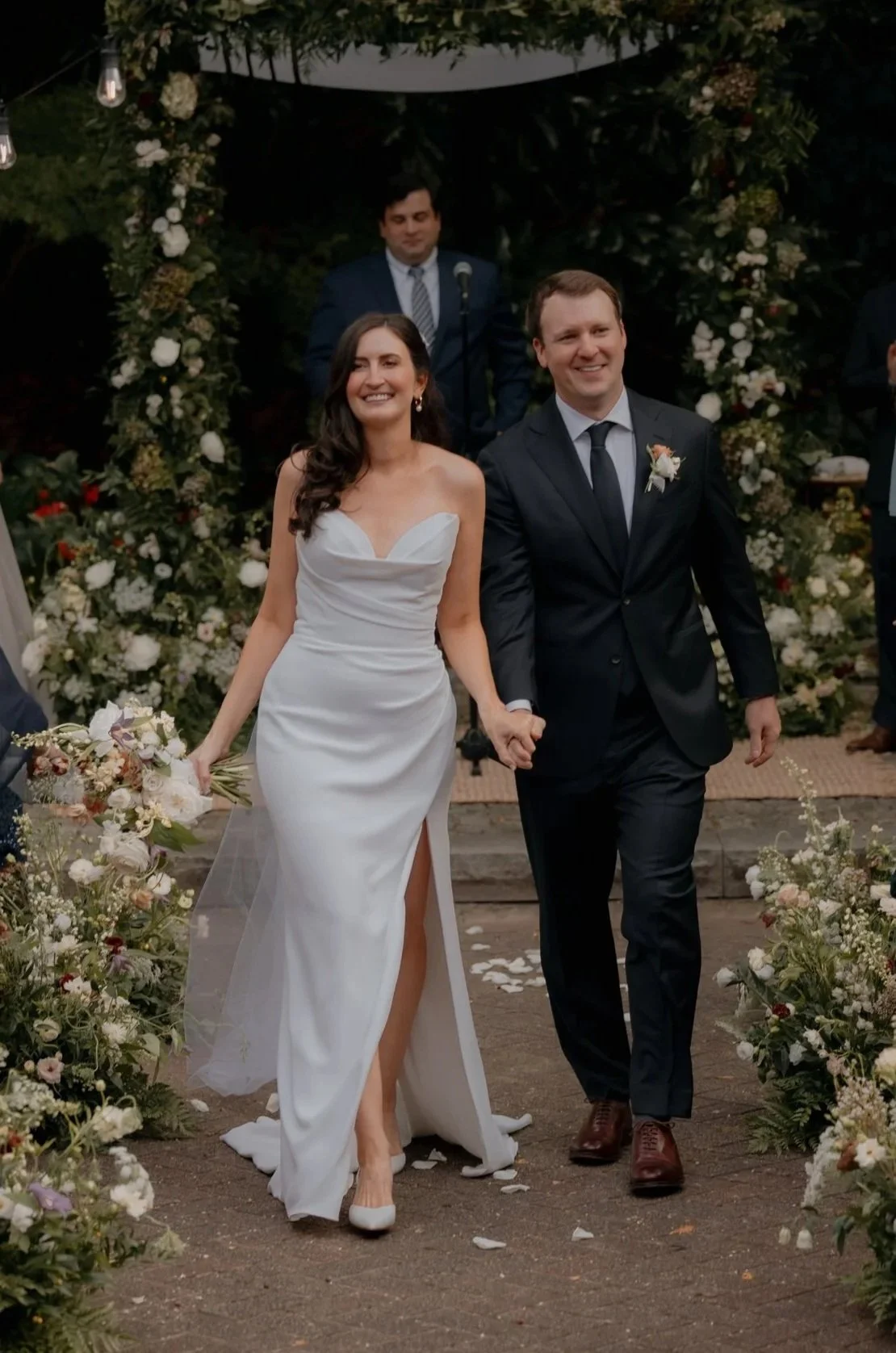 A newlywed couple walking down the aisle, holding hands, surrounded by floral arrangements, with a man in a suit standing in the background at a wedding ceremony.
