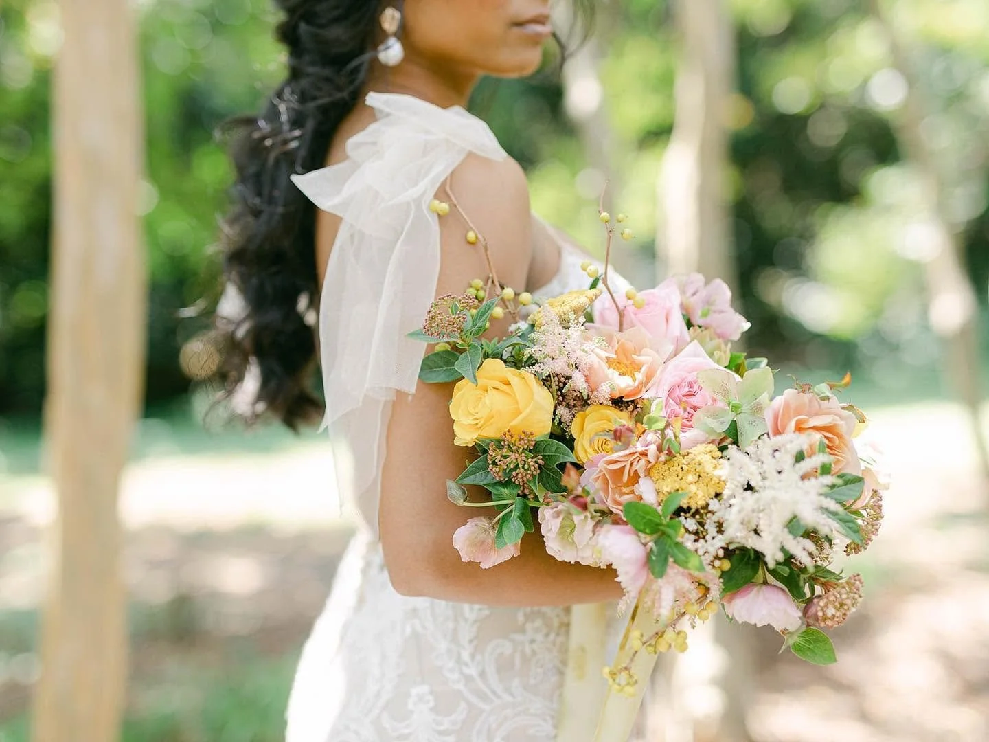 A bride holding a large bouquet of pink, yellow, and white flowers with greenery, outdoors in a sunny, wooded area.