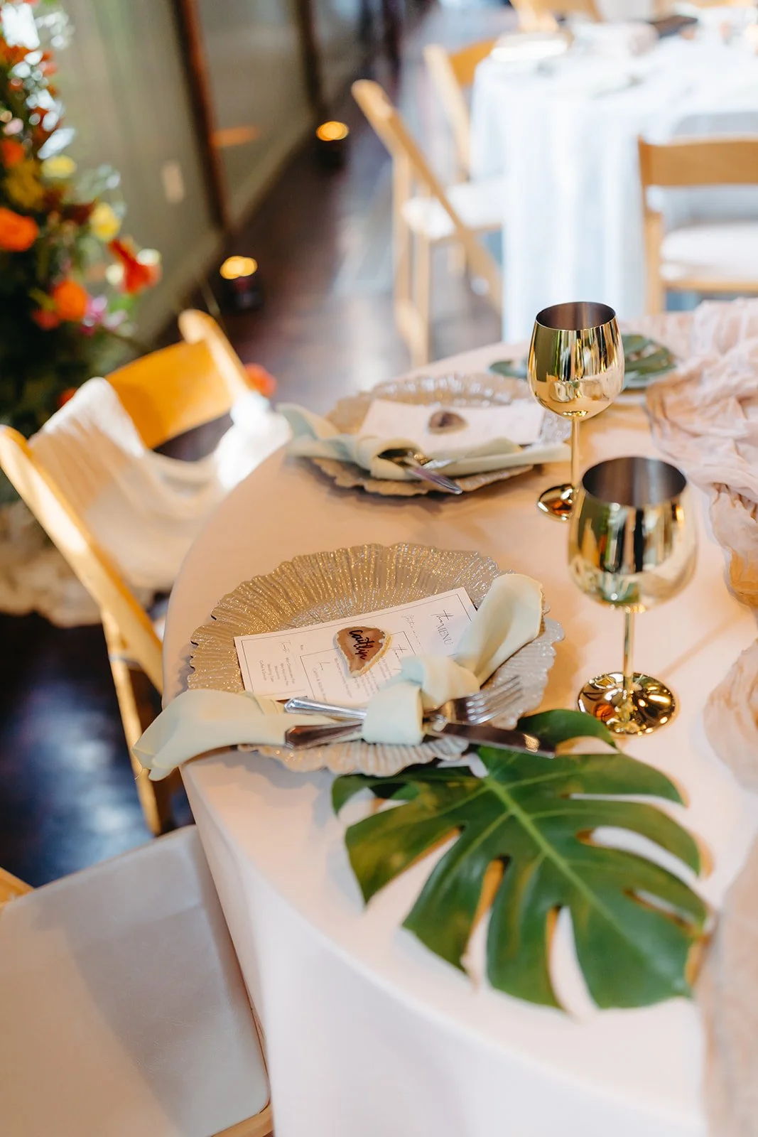 Elegant dining table set with gold glasses, tropical leaves, and a holiday-themed menu with a heart-shaped cookie, amidst candles and flowers.