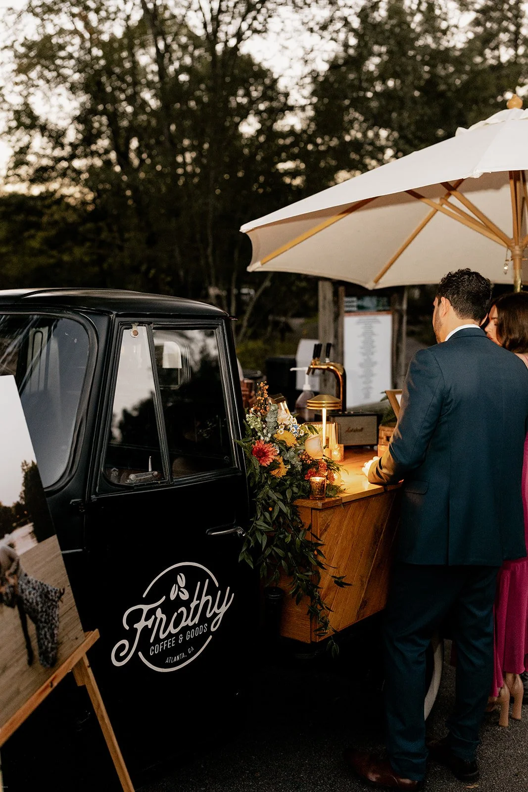 A black coffee and goods cart with a floral decoration, serving drinks at an outdoor event. People in formal attire are standing under a large beige umbrella, with trees and a menu board in the background.