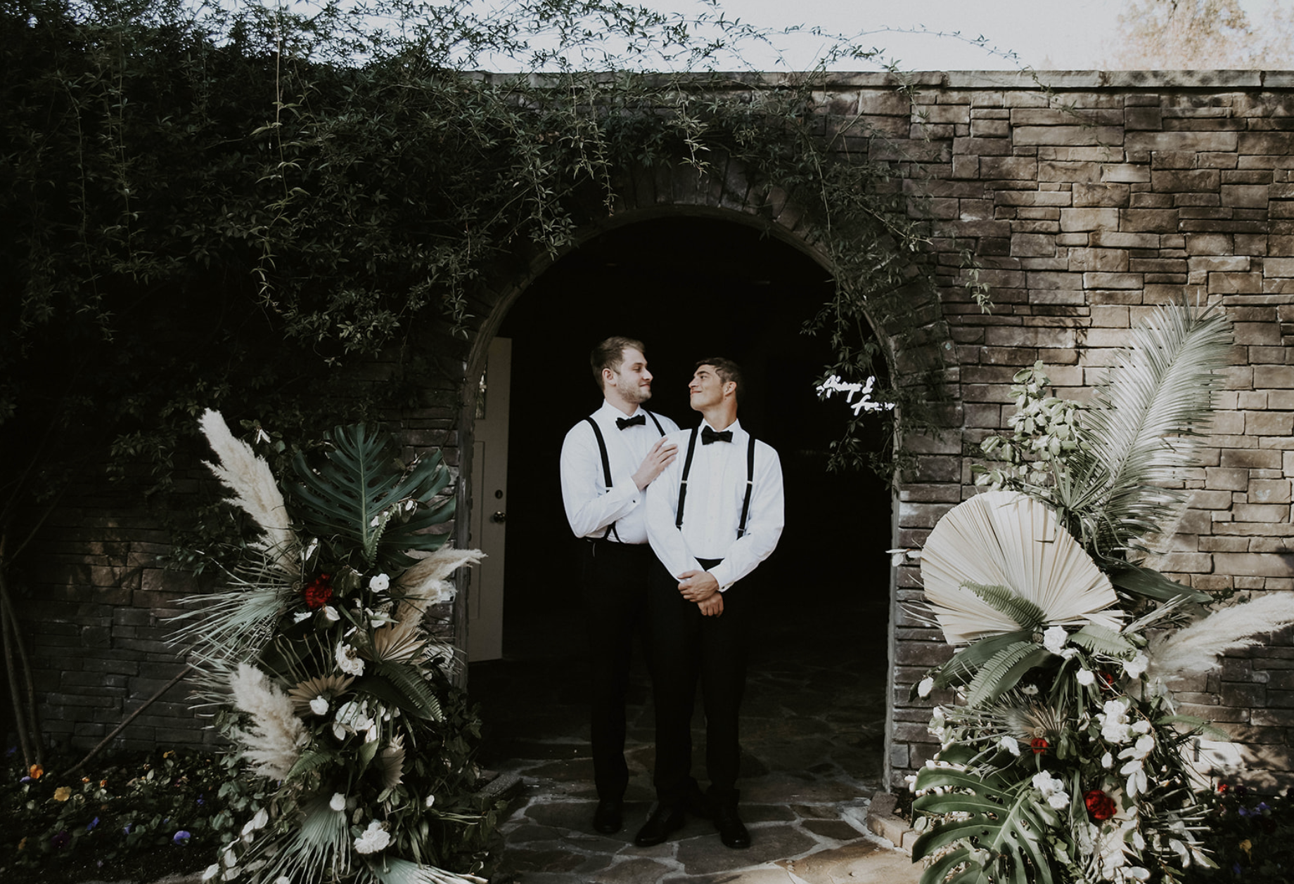 Two men in formal attire, with white shirts and black suspenders, standing close together under a stone archway decorated with foliage and flowers, smiling at each other during a wedding or celebration.