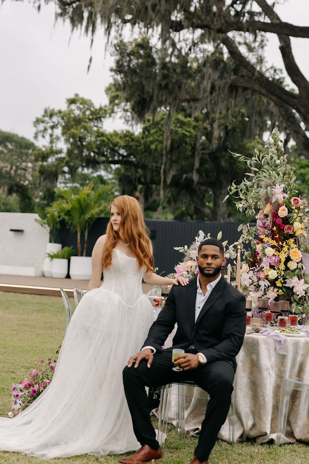 A bride in a wedding dress and a groom in a black suit are at an outdoor wedding reception with a decorated table and floral arrangements. The bride is standing, and the groom is sitting holding a drink.