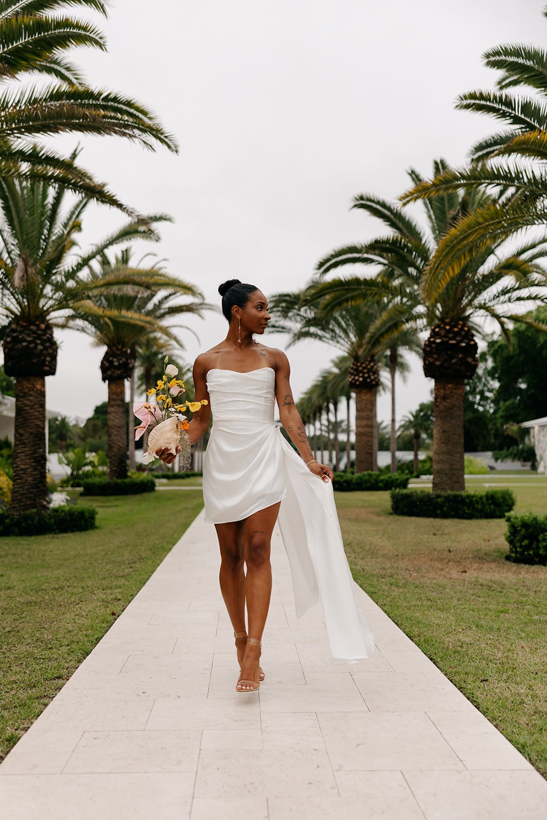 A woman in a white dress holding a bouquet walking on a stone pathway lined with palm trees on a cloudy day.