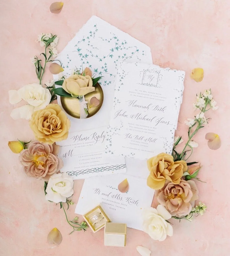 Wedding invitations surrounded by floral arrangements and a small jewelry box on a pink background.