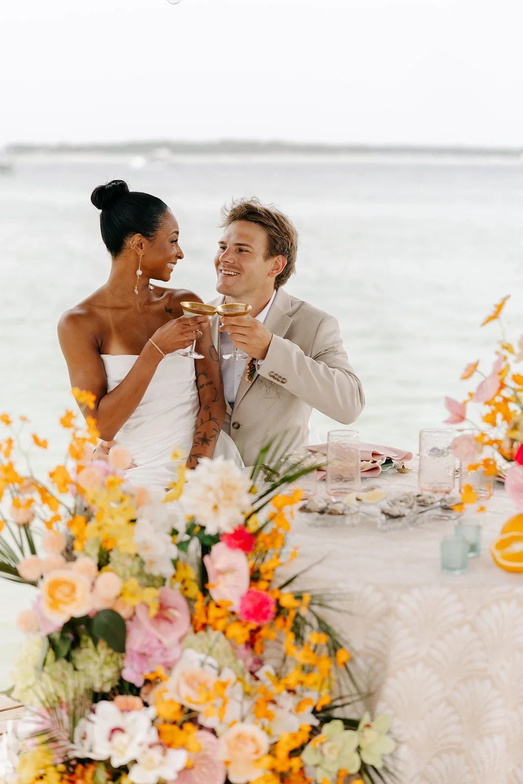 A newlywed couple celebrating with drinks at a wedding reception by the water, decorated with colorful flowers and table settings.