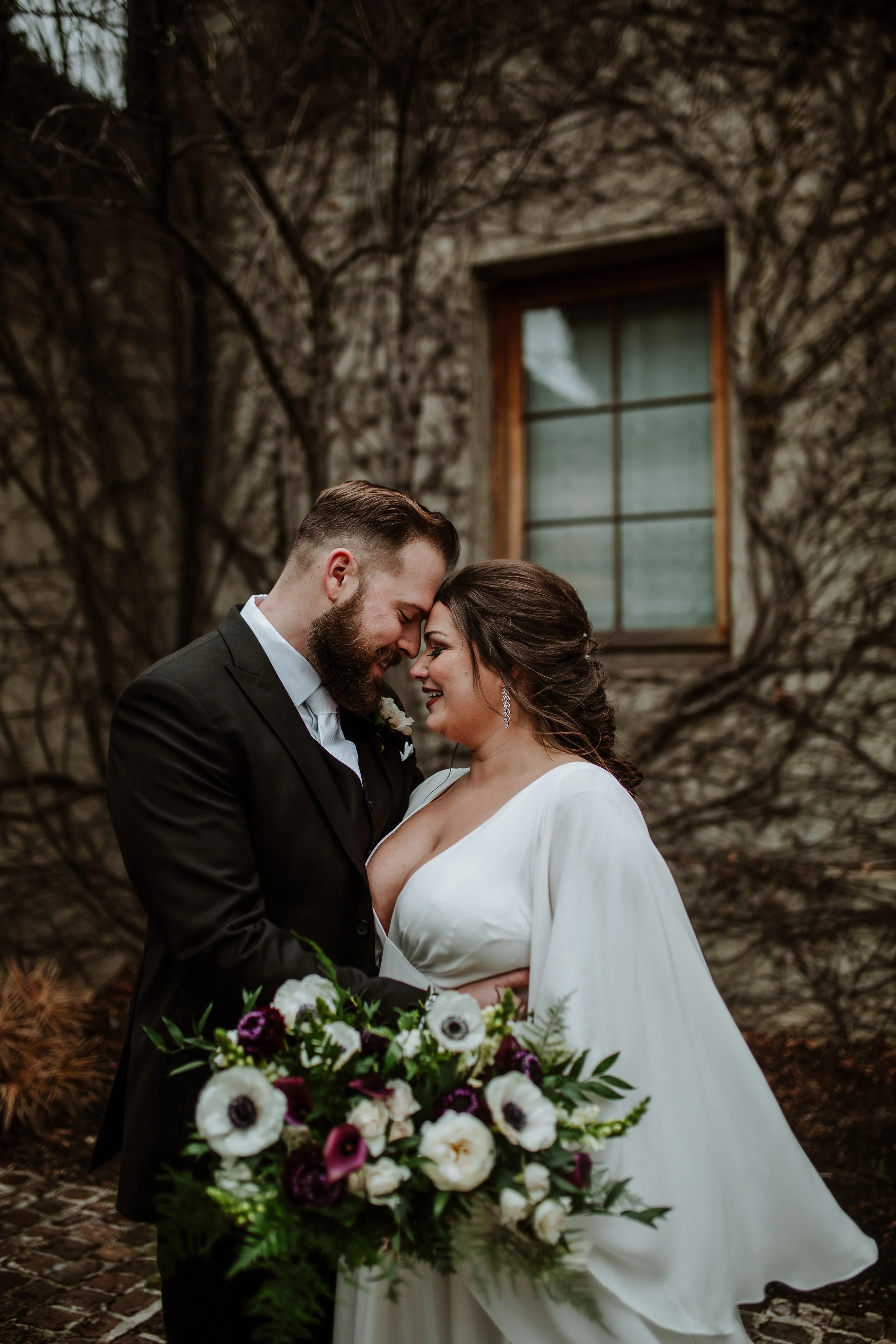 A bride and groom share a close moment, smiling and touching foreheads, holding a bouquet of white, purple, and dark red flowers outdoors with a brick pathway and a large window in the background.