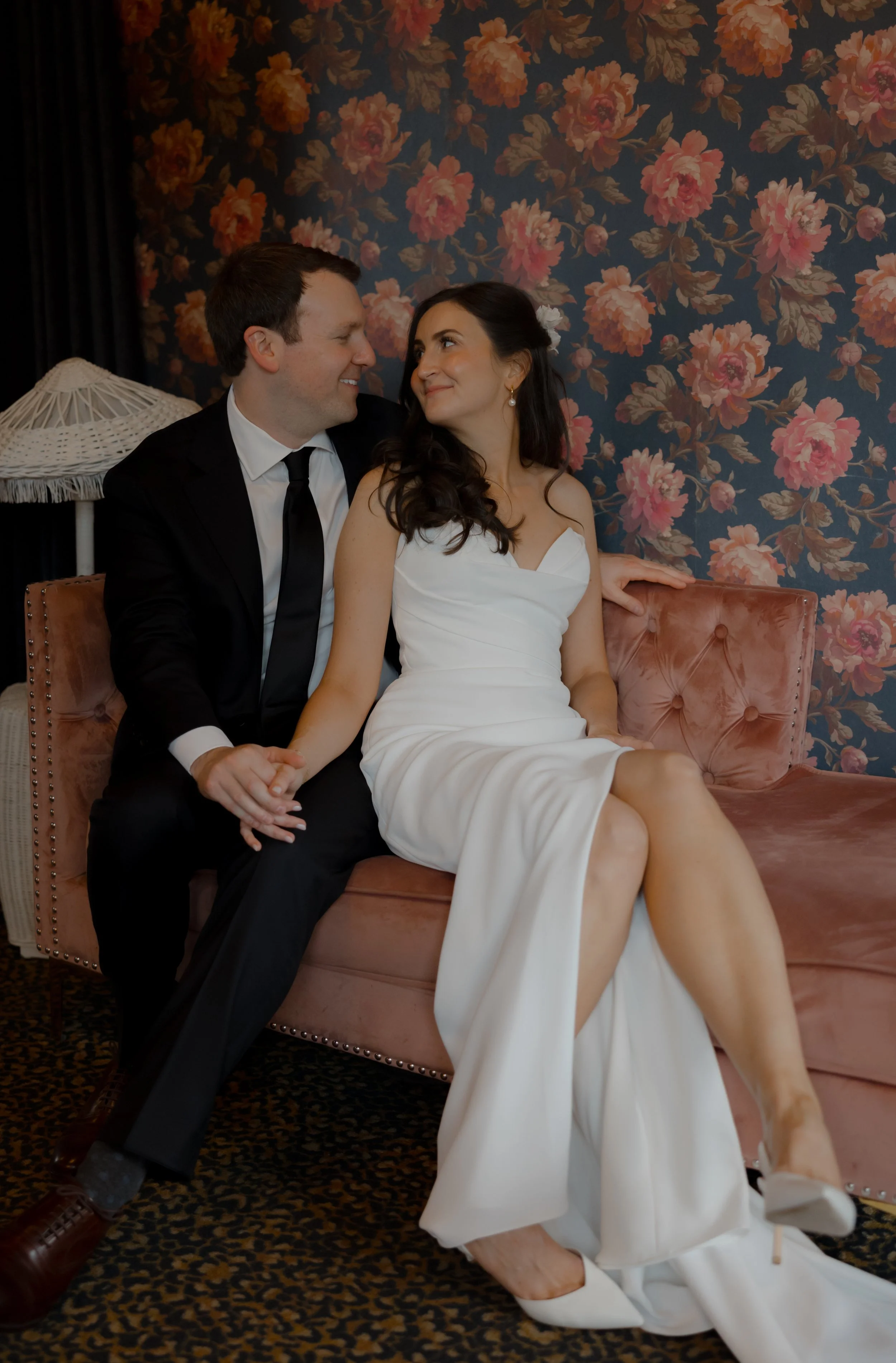 A bride and groom sitting on a pink velvet couch, gazing into each other's eyes with a floral wallpaper background. The bride is wearing a white strapless wedding dress and high heels, while the groom is dressed in a black suit with a white shirt and black tie.
