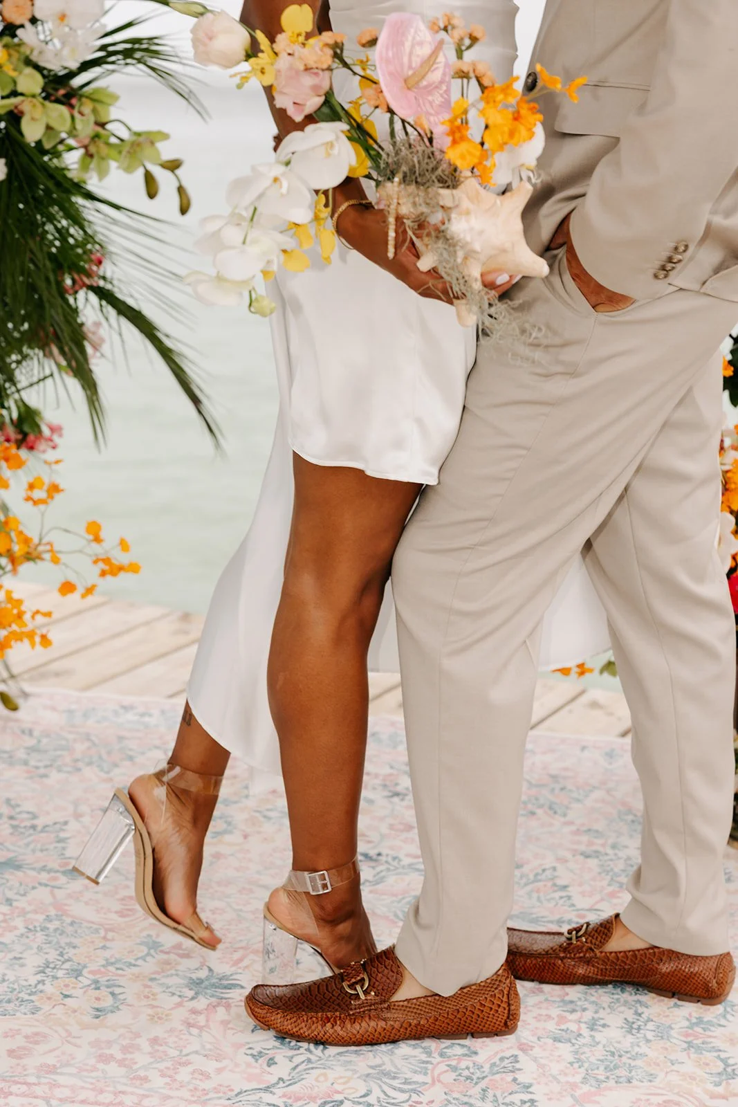 Close-up of a couple standing on a patterned rug, holding a bouquet of flowers, with the woman wearing high-heeled sandals and the man in brown woven loafers, with a floral background and water in the distance.