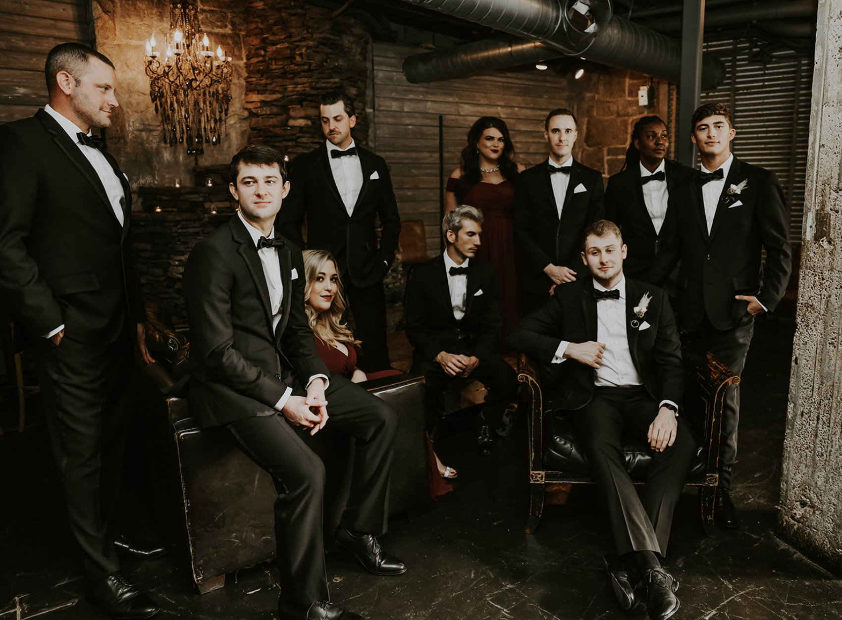 A group of eleven young adults in formal tuxedos and evening dresses in a dimly lit, rustic room with exposed brick and wooden walls, some sitting and some standing, looking at the camera.