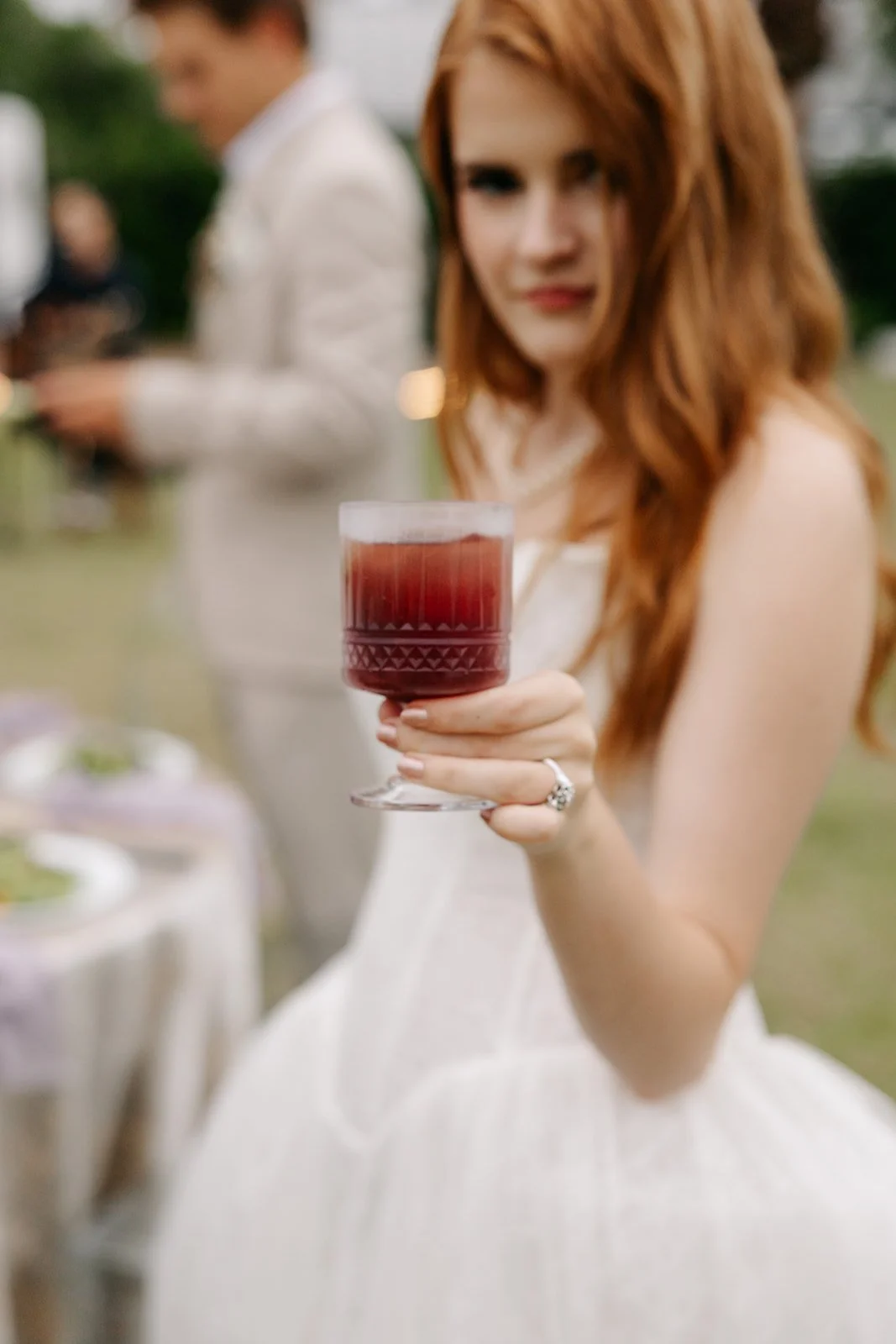 A woman with red hair in a white dress holds a glass of red wine toward the camera, with a blurred outdoor gathering in the background.