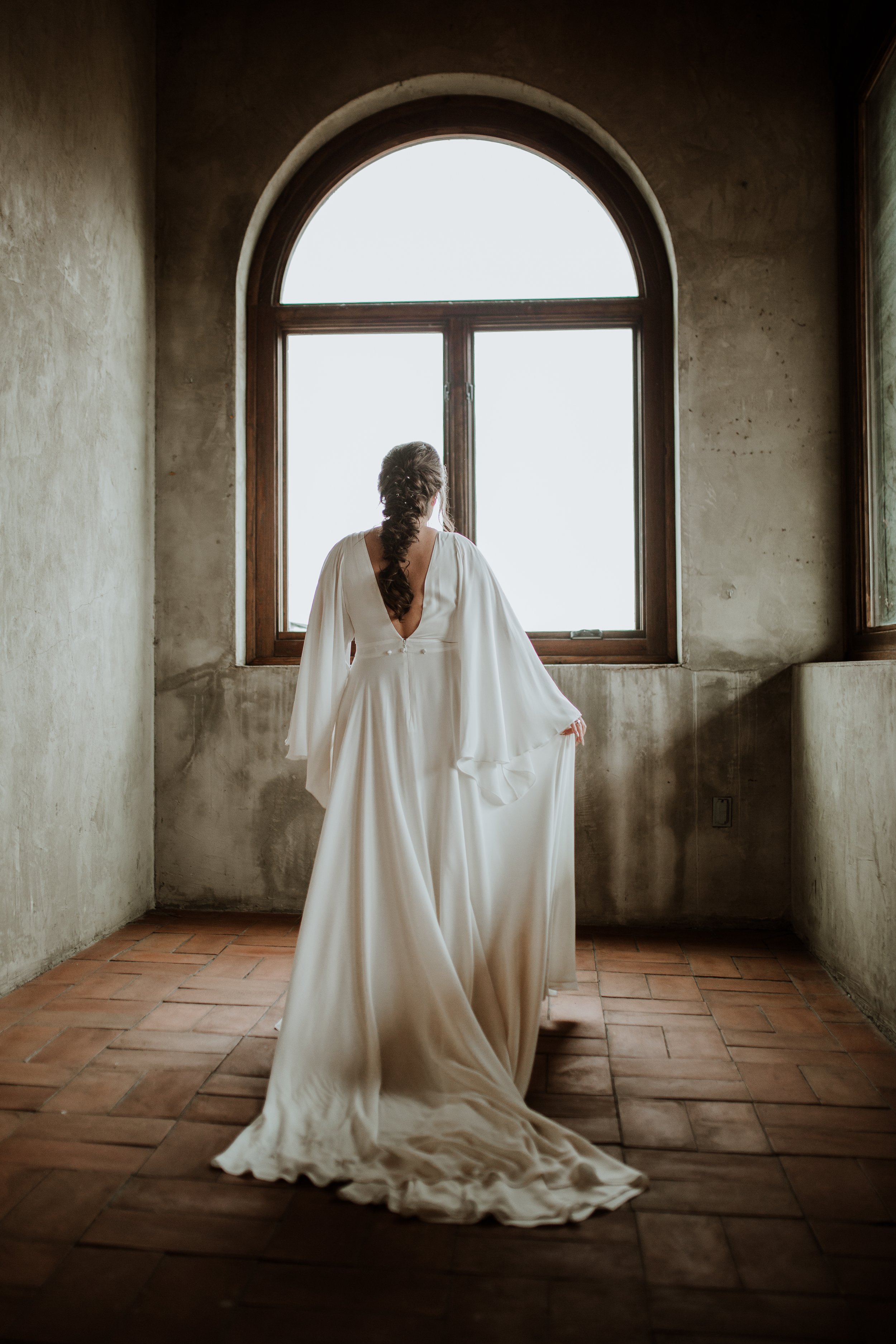 A woman in a long white dress with a deep back, standing in front of a large arched window, looking outside in a room with rough textured beige walls and terracotta tiled floor.
