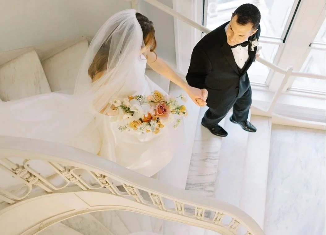 A bride in a wedding dress holding a bouquet looking down a staircase, holding hands with a groom in a tuxedo.
