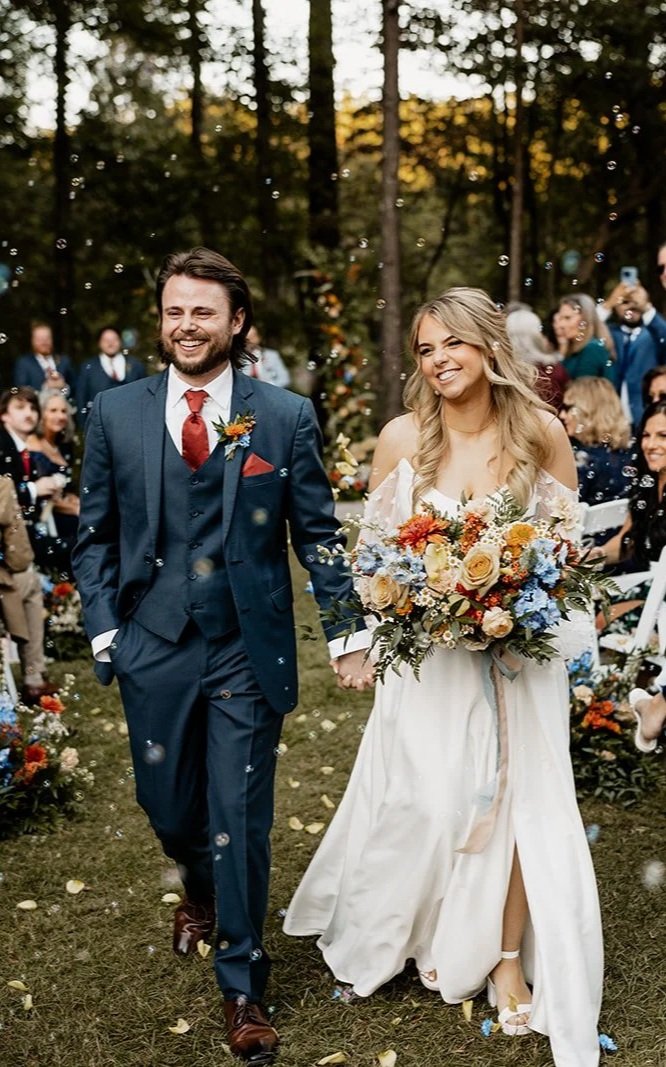 A happy bride and groom walking hand-in-hand outdoors during a wedding ceremony, surrounded by guests and trees, with colorful floral arrangements and confetti falling.