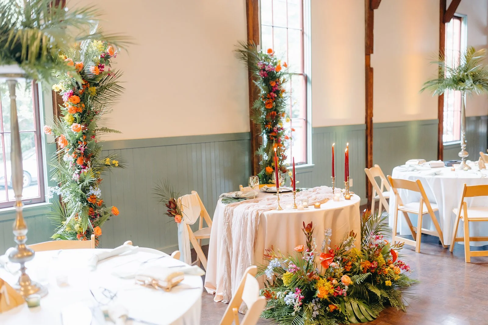 Elegant event setup with round tables draped in white tablecloths, decorated with tall candleholders with red candles, floral arrangements, and table settings, set in a room with large windows, wooden beams, and floral arch decor.