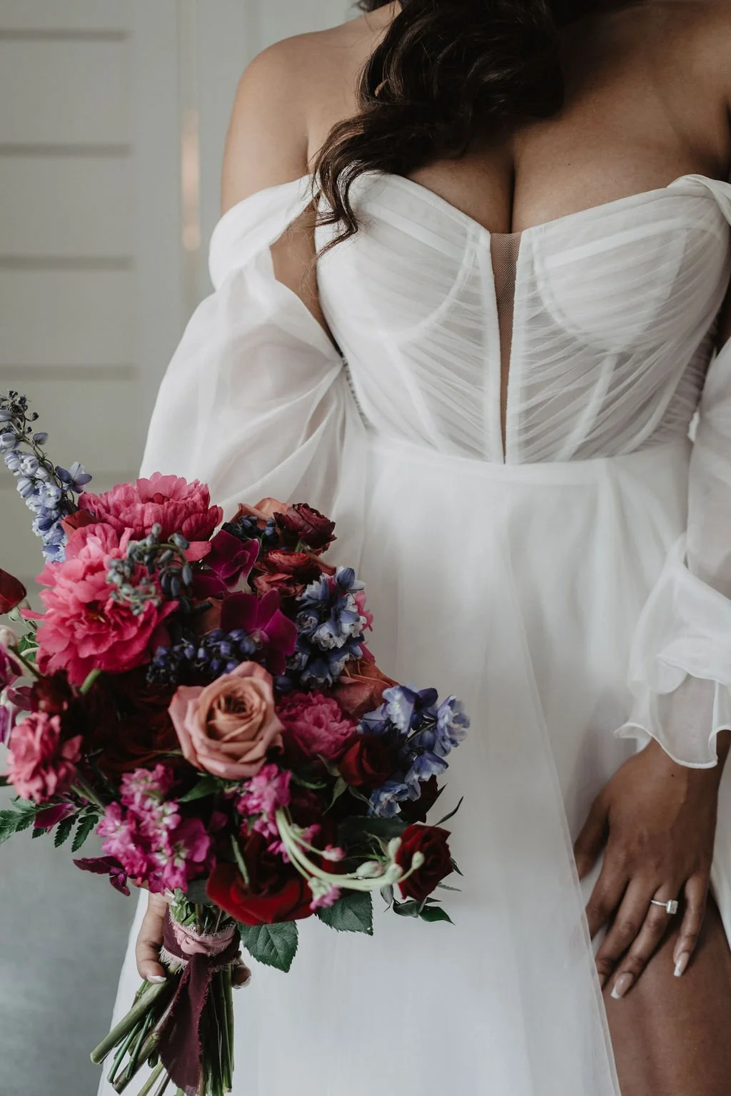 A bride holding a bouquet of pink, purple, and red flowers, wearing a white wedding dress with a fitted bodice and off-the-shoulder sleeves.
