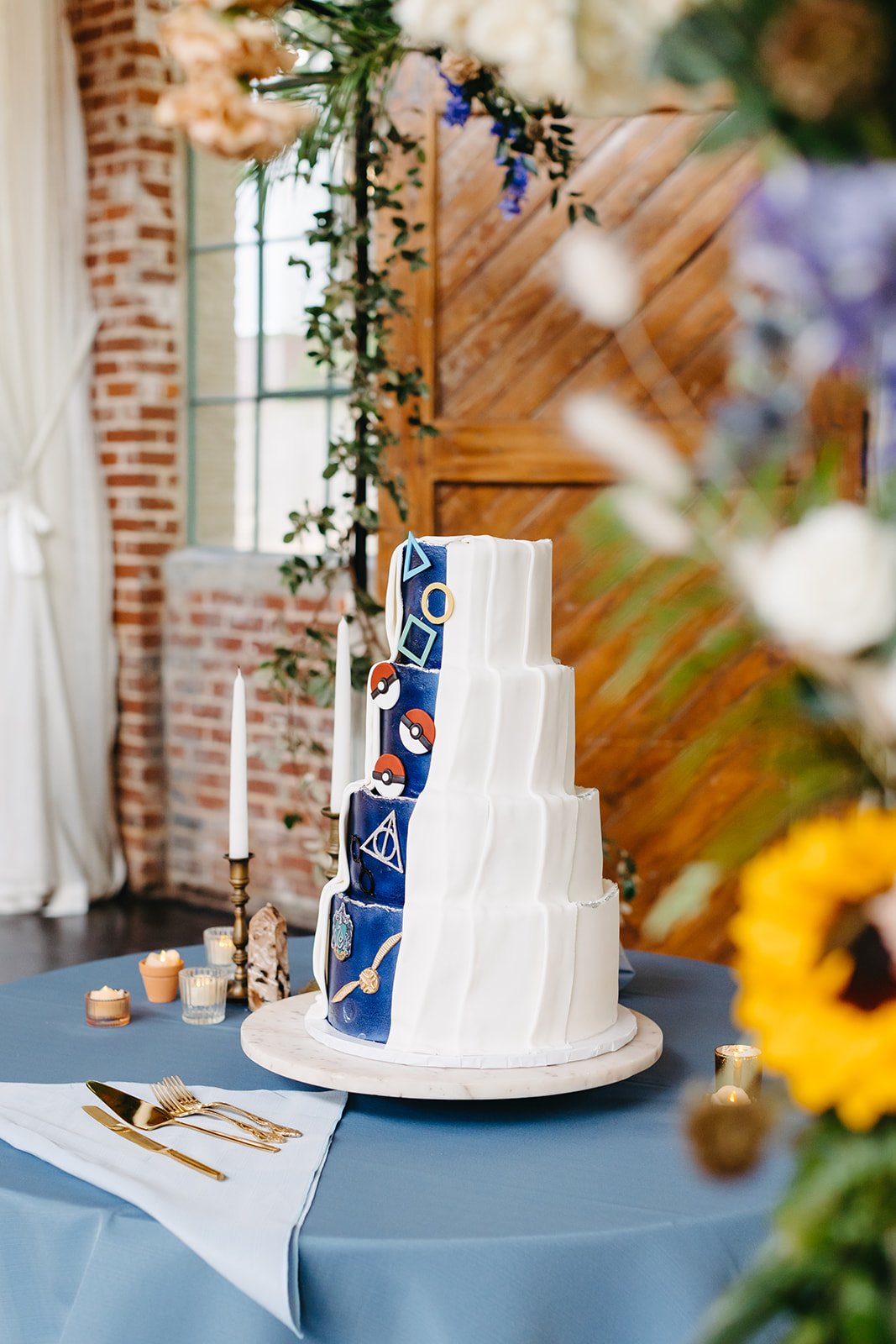 A tiered white wedding cake with a blue ribbon featuring Pokémon symbols, placed on a table with a blue and white tablecloth, surrounded by candles and cutlery, in a rustic indoor setting with brick and wooden accents.