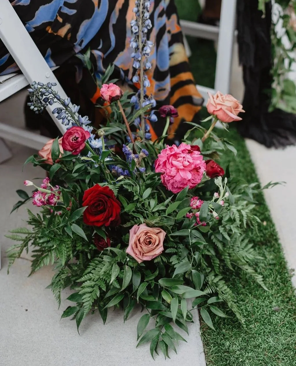 A colorful bouquet of pink, red, and peach roses, peonies, and purple flowers arranged with green foliage on a table outdoors.