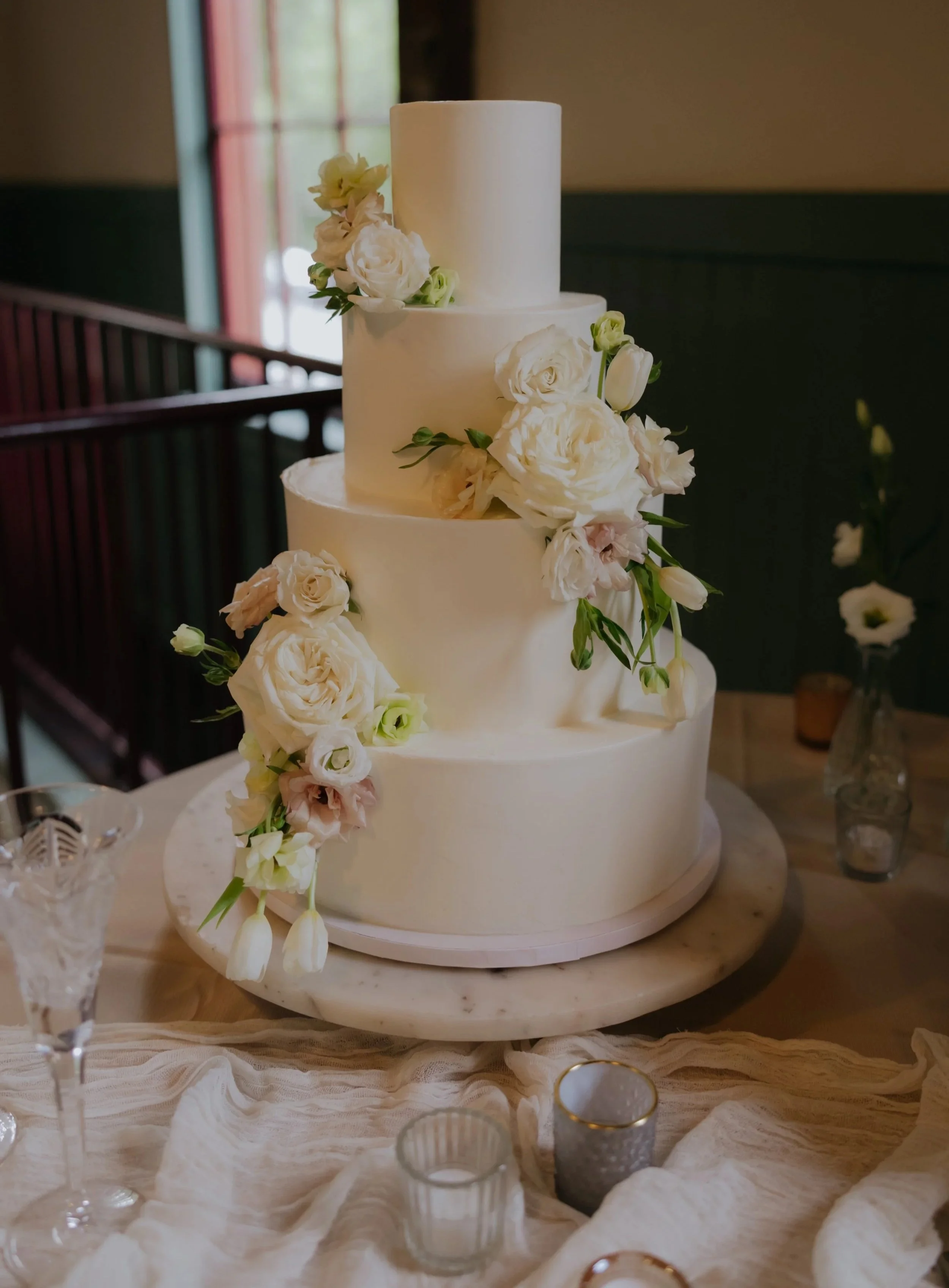 Three-tier white wedding cake decorated with white flowers, placed on a round marble cake stand, surrounded by candles and glassware on a draped table.