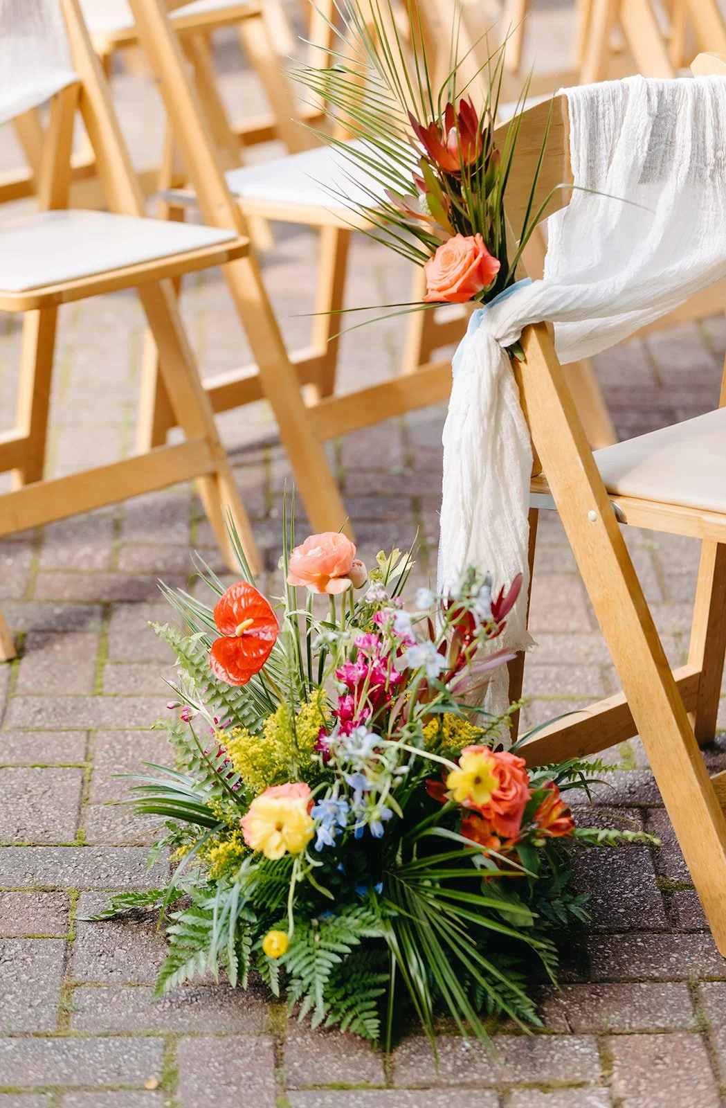 Decorated wooden chairs arranged outdoors with floral arrangements featuring pink, orange, and yellow flowers, and a white fabric draped over one chair for a wedding or event.