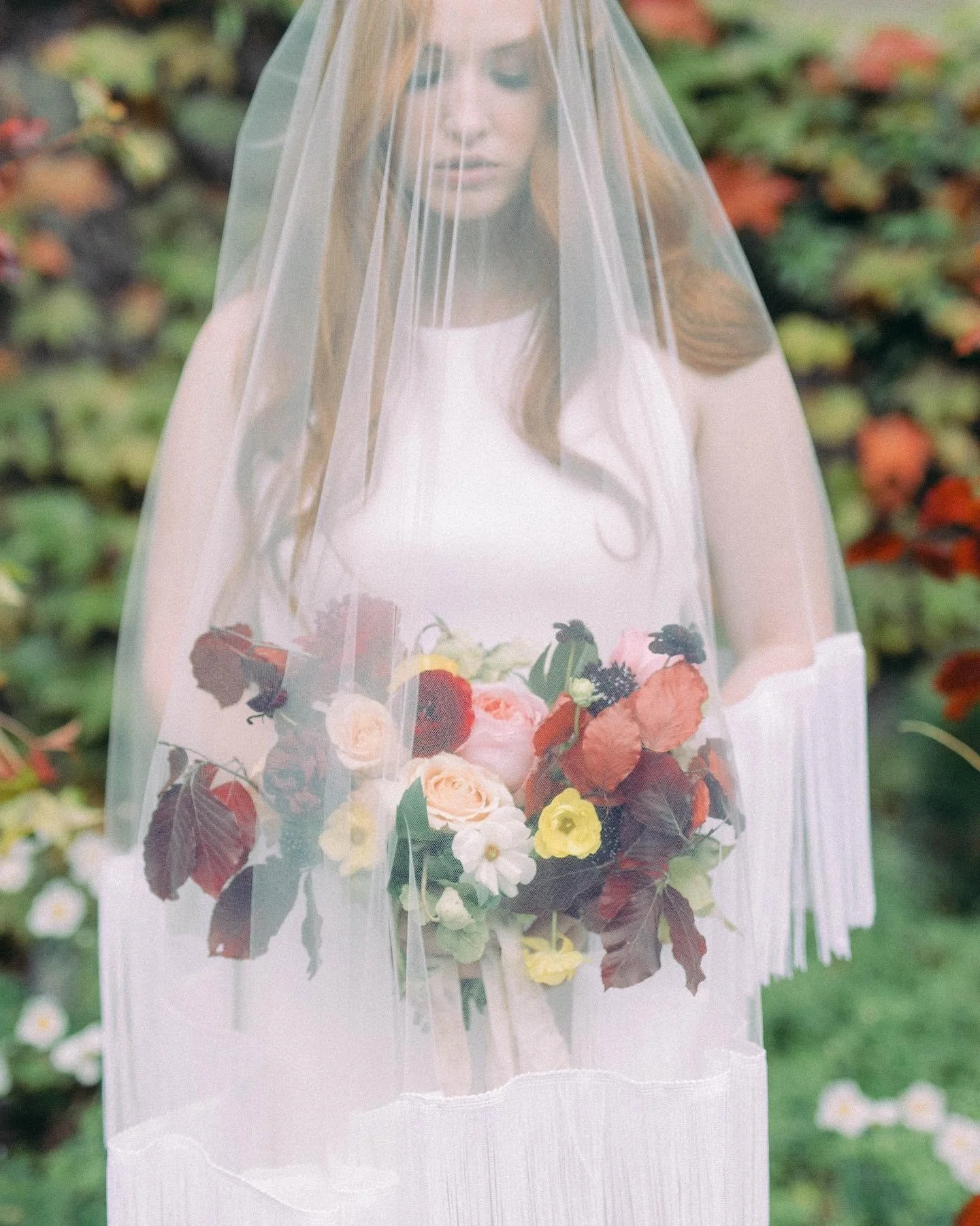 A woman with red hair standing outdoors behind a sheer white veil, holding a colorful bouquet of flowers, with blurred greenery and flowers in the background.