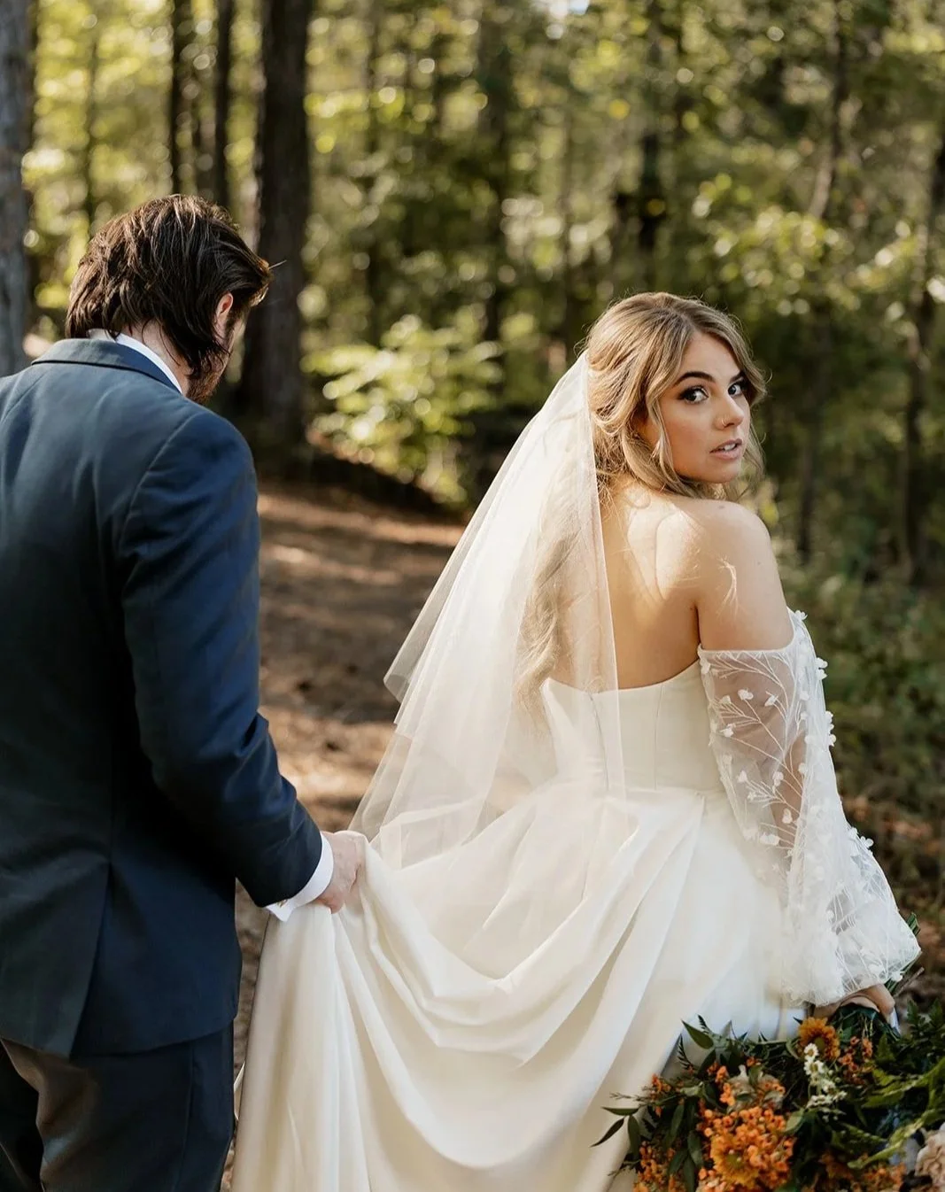 Bride in an off-shoulder wedding dress with a veil holding a bouquet, standing in a forest, with a man in a suit holding her dress.