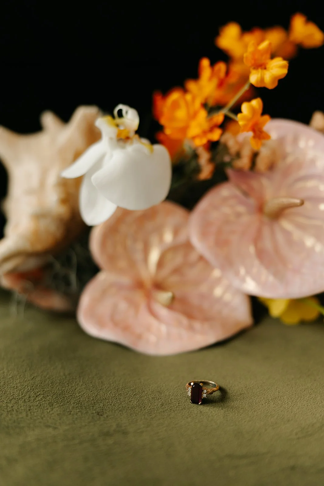 A gold ring with a large rectangular dark red gemstone, possibly garnet or ruby, sits on a green surface. In the background, there are seashells, orange flowers, and a white decorative object, with a dark backdrop.