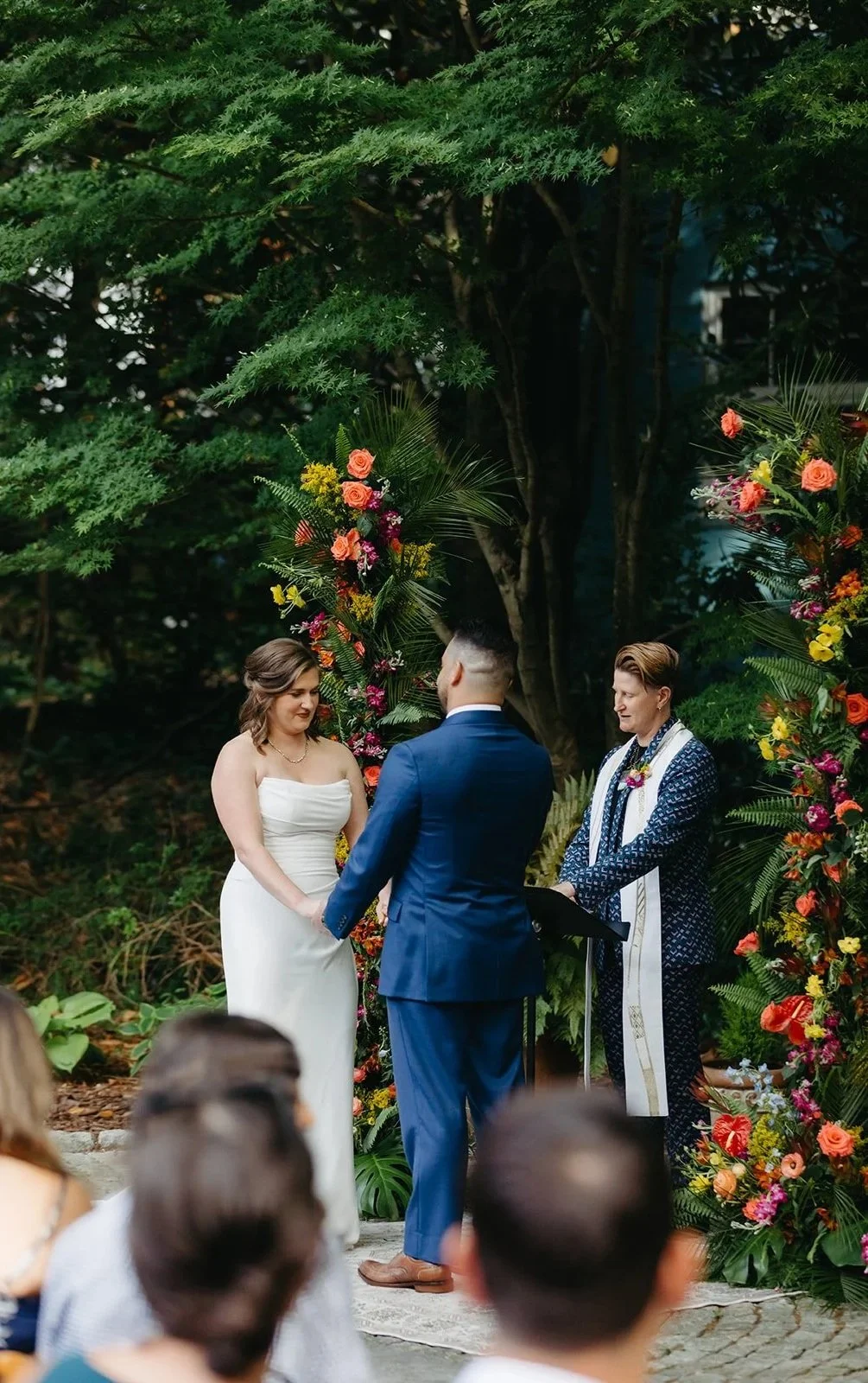 A couple holding hands during their outdoor wedding ceremony, with an officiant standing beside them in front of a floral arch, surrounded by greenery.