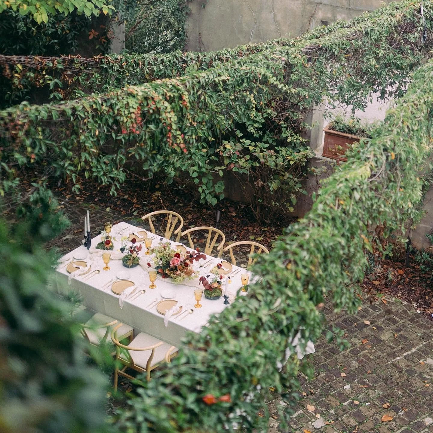 An outdoor dining table set for a formal event, decorated with flowers, candles, and elegant tableware, surrounded by greenery and cobblestone ground.
