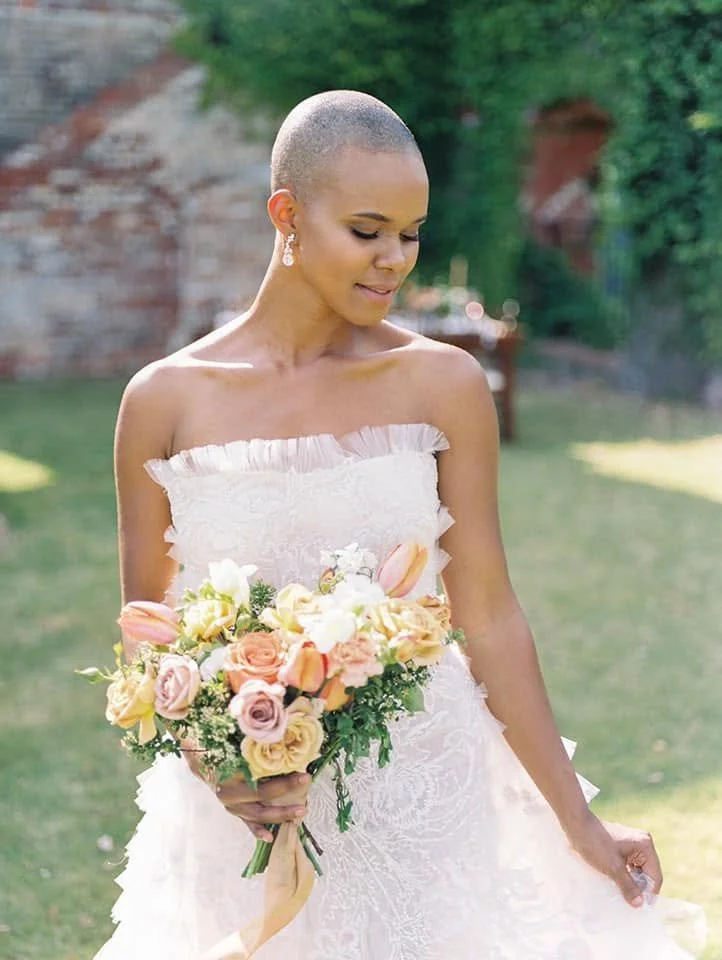A woman in a white wedding dress holding a bouquet of pastel-colored roses and lilies outdoors, with green trees and a brick wall in the background.