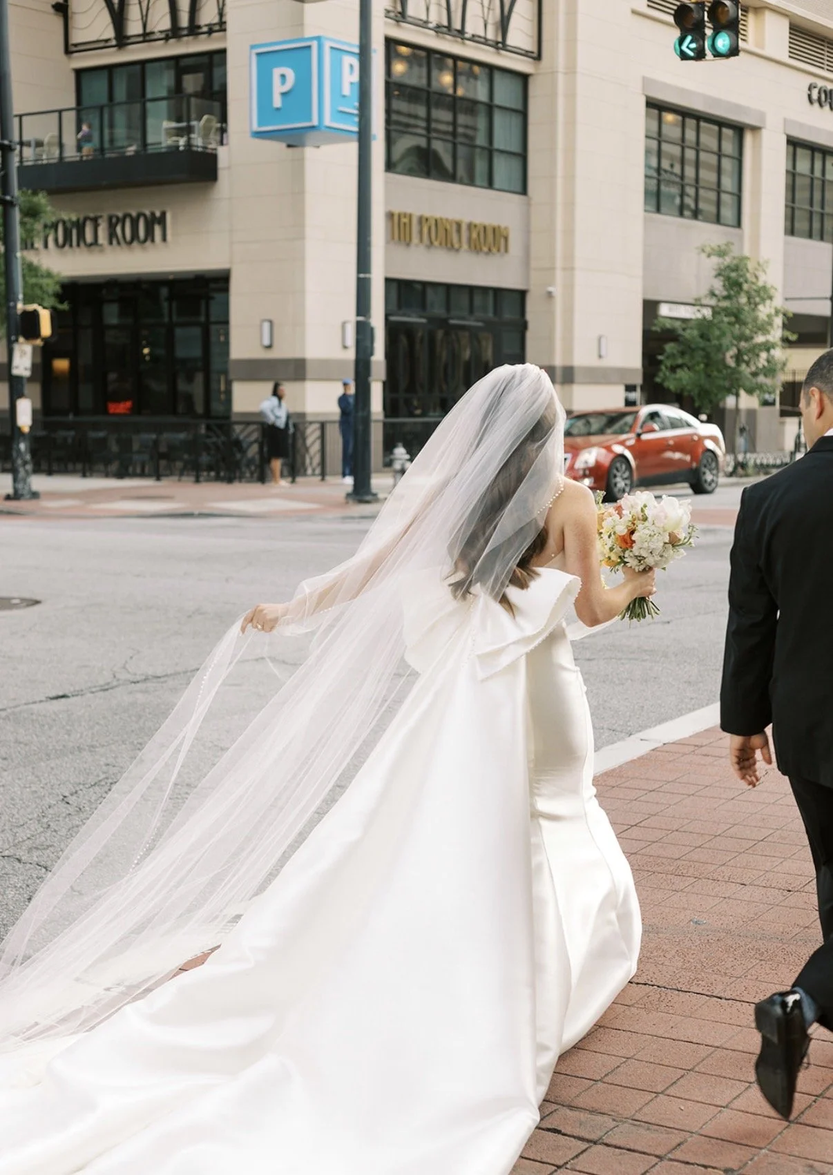 A bride in a white wedding dress with a long train and veil, holding a bouquet of flowers, walking down the street with a groom in a black suit. Buildings and cars are visible in the background.