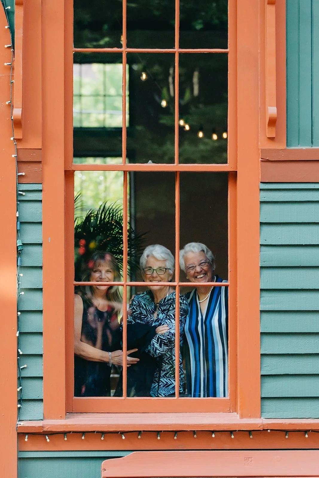 Three women standing behind a window with orange trim, smiling at the camera.