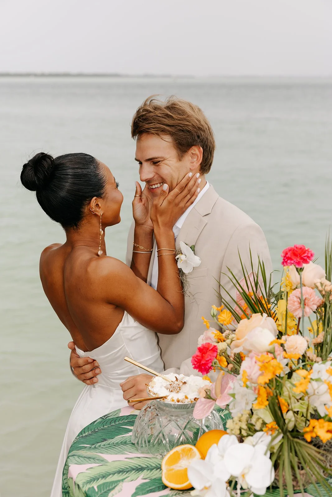 A couple, dressed in wedding attire, sharing an intimate moment on a beach with water in the background. The woman has dark hair in a bun and is touching the man's face, who has brown hair and a light-colored suit with a white boutonniere. There is a table with a floral arrangement, oranges, and dessert in front of them.