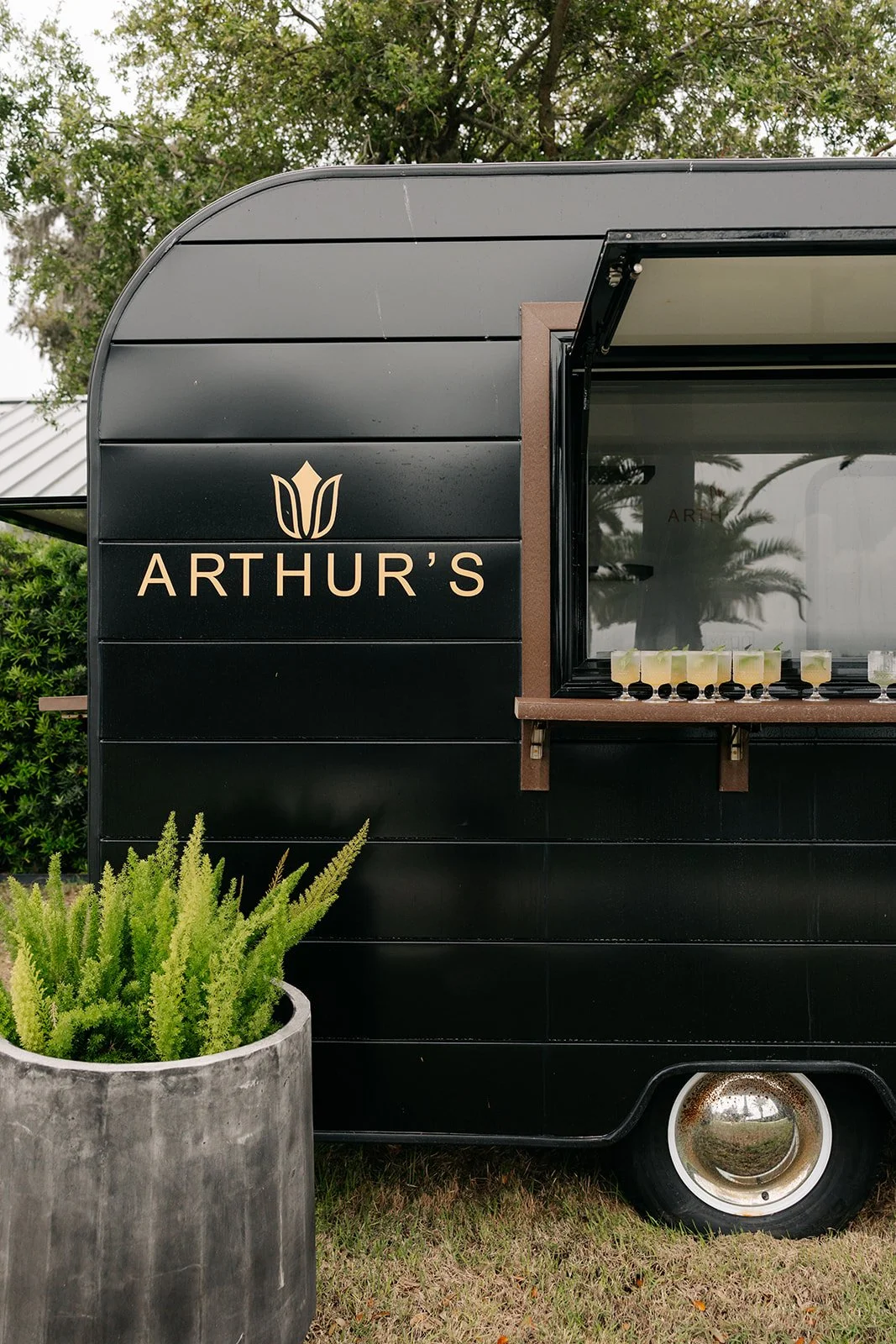 Black food truck with 'Arthur's' sign and small window serving drinks, with a large potted green plant in front.