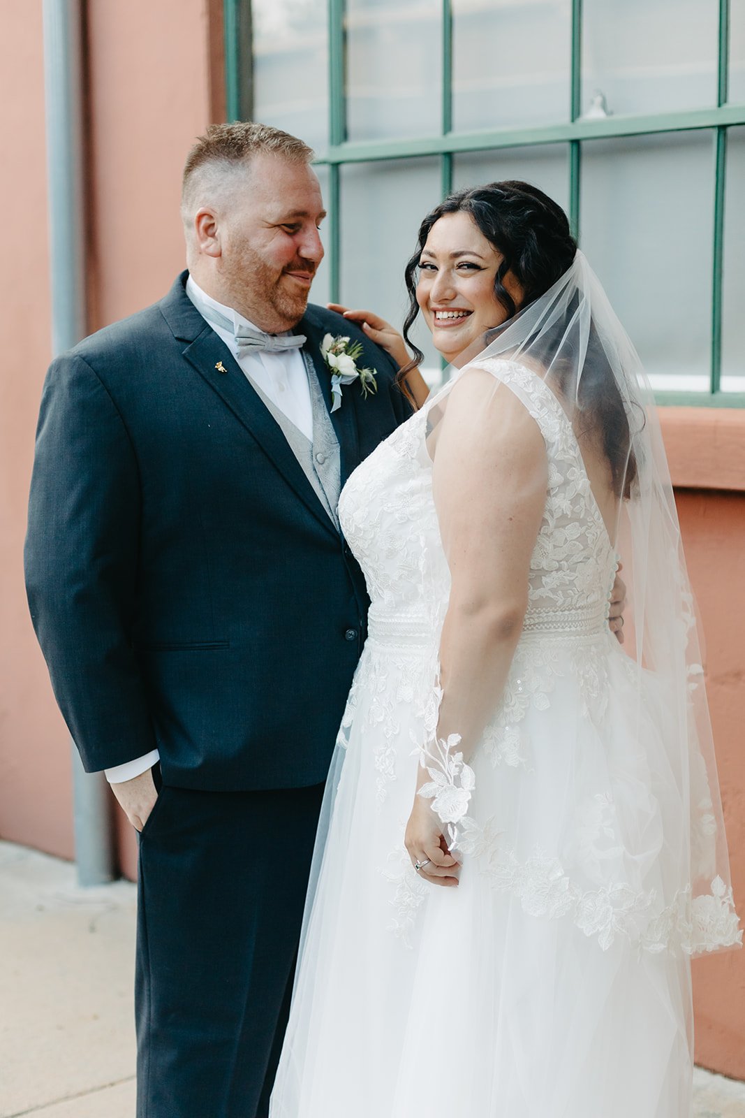 A bride and groom on their wedding day, smiling and looking happy, standing outdoors in front of a building with large green windows.
