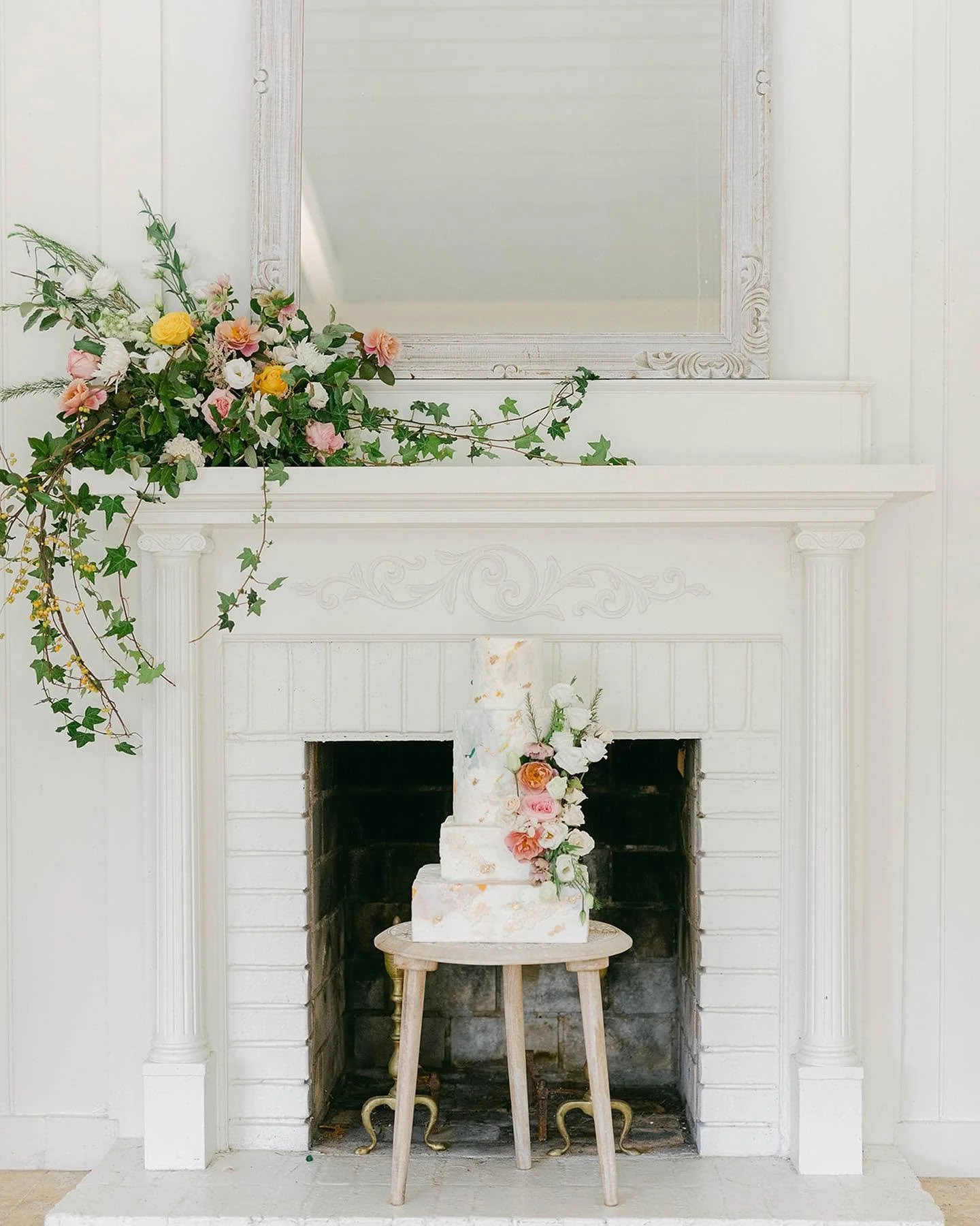 A white fireplace with decorative molding, a large mirror above it, and a floral arrangement including pink, yellow, and white flowers on the left side and a cake with flowers on the right, placed on a small stool in front of the fireplace.