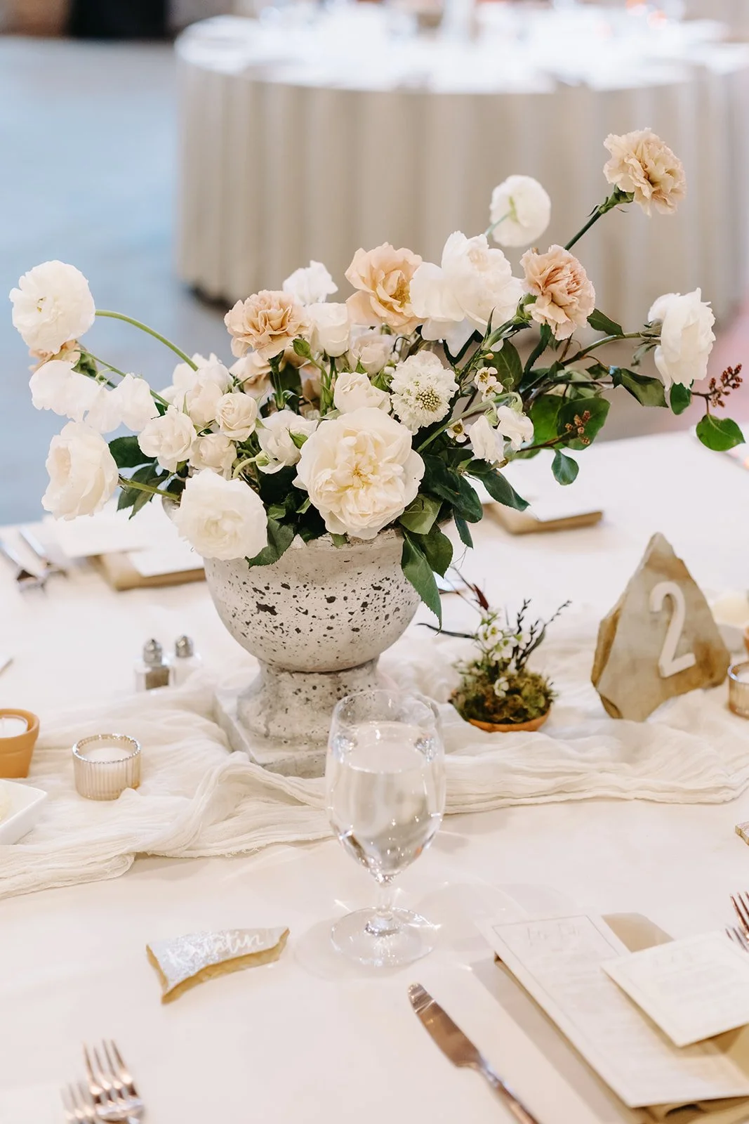 A table centerpiece featuring a white and blush flower arrangement in a speckled stone vase, with a glass of water and table settings, at a formal event or wedding reception.