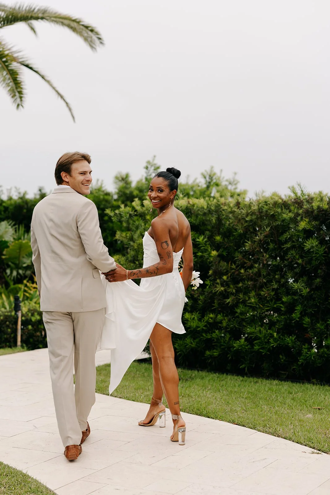 A newlywed couple holding hands and smiling outdoors on a cloudy day, with greenery in the background, the woman wearing a white strapless wedding dress and high heels, the man in a light-colored suit.